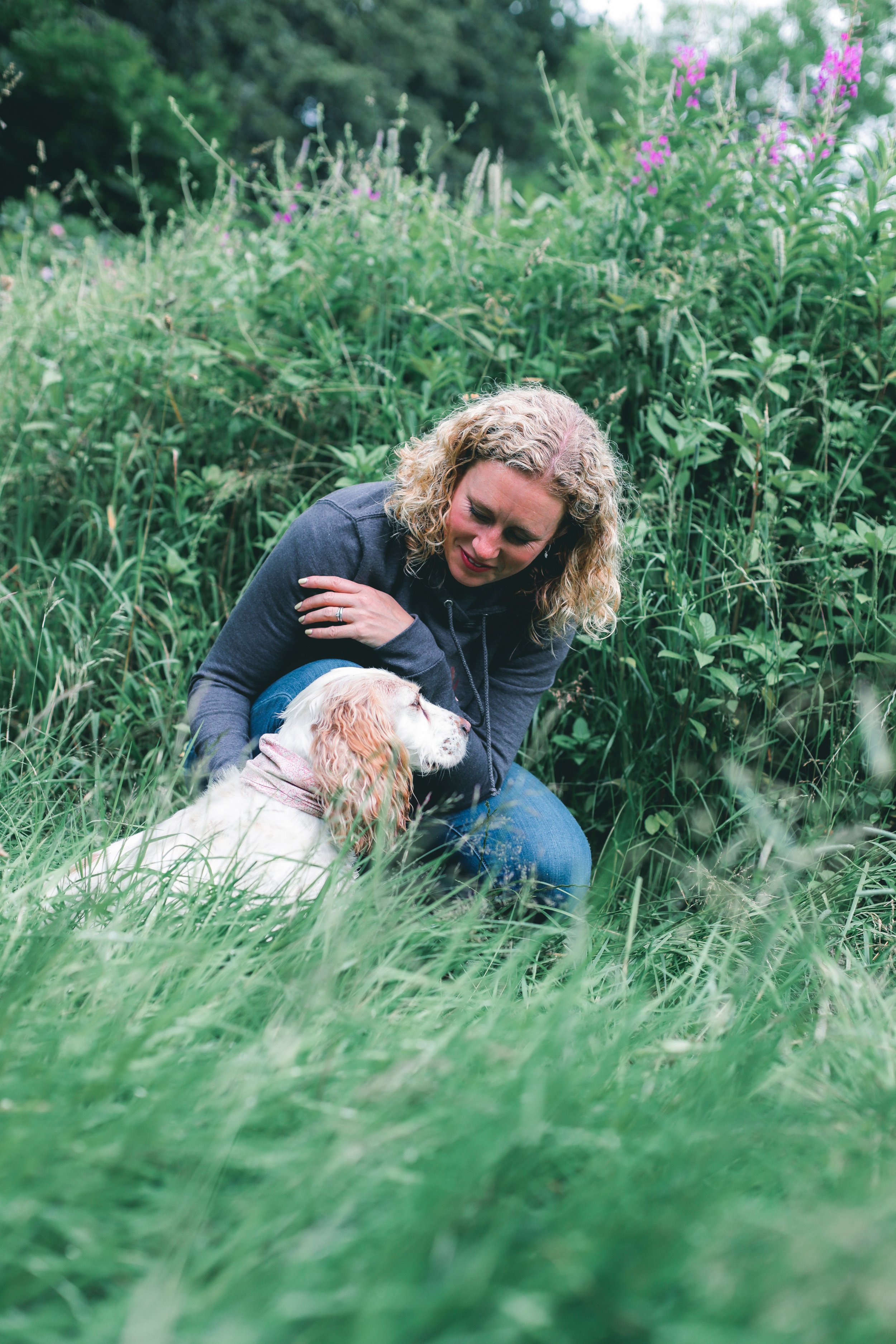 Woman in gray hoodie sitting in tall grass holding a white and brown dog in a lush, green outdoor setting.