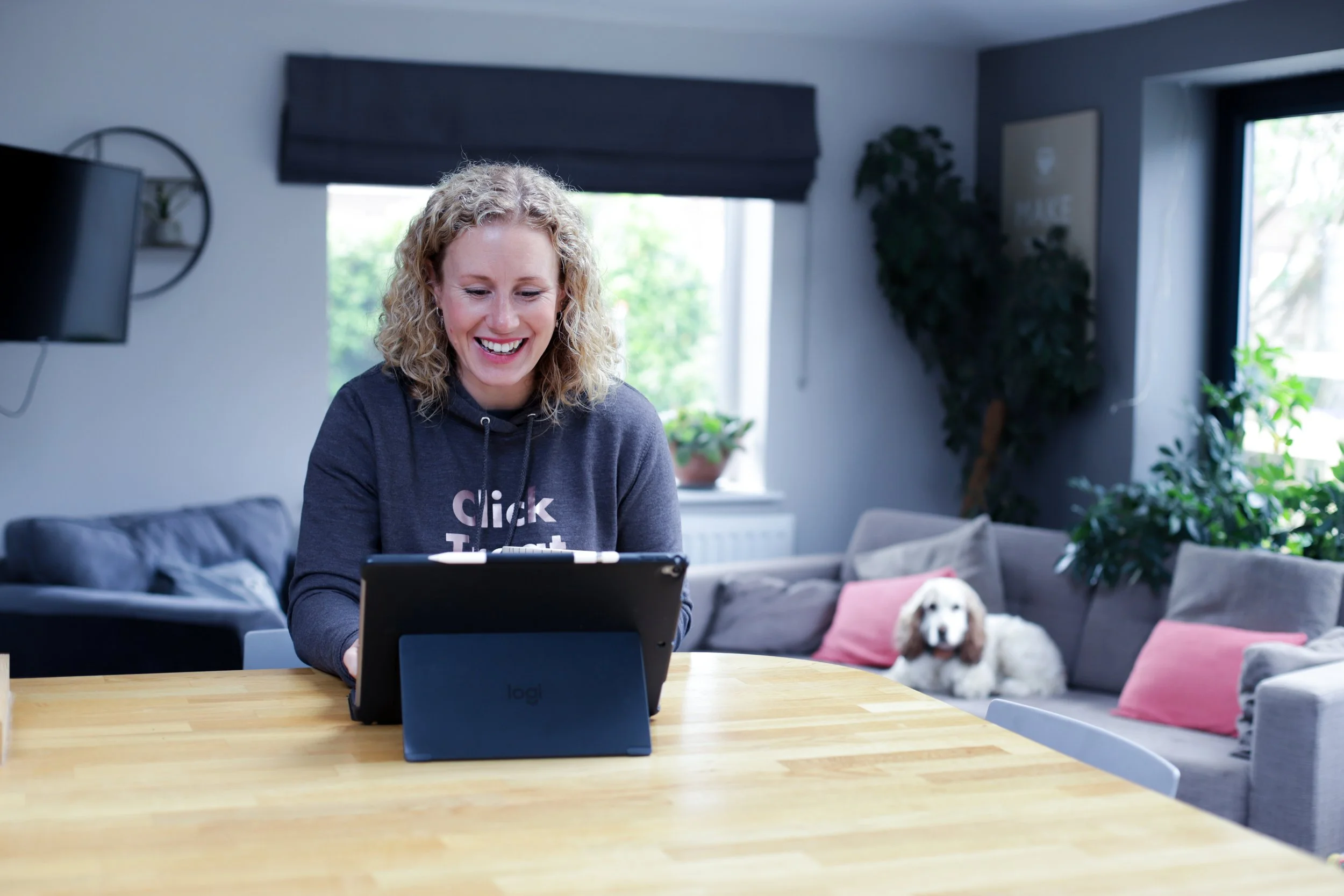 A woman with curly blonde hair smiling while looking at a tablet on a wooden table in a bright living room with large windows, a gray sofa with pink cushions, and houseplants, with a small white and brown dog in the background.