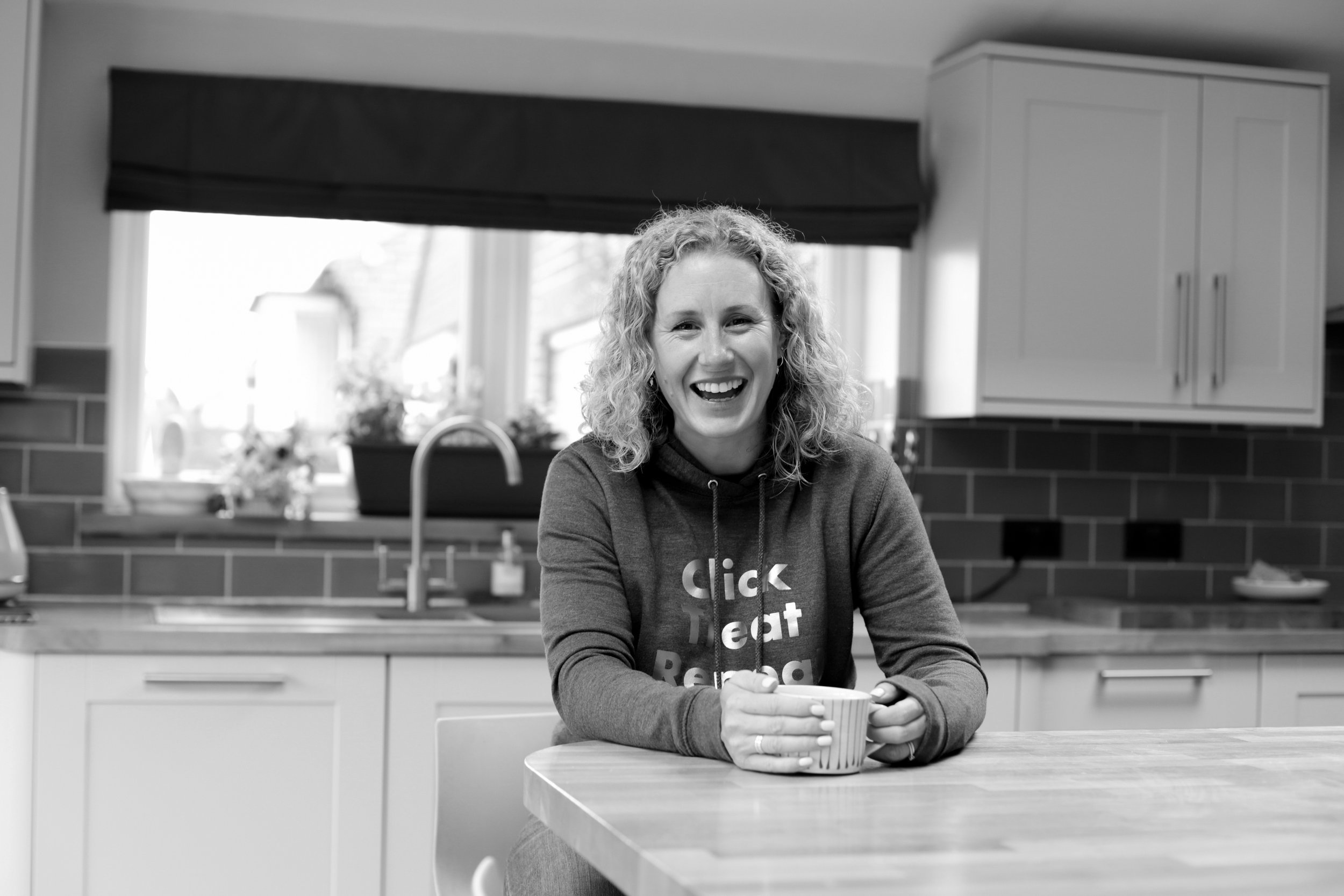 A woman with curly hair smiling and sitting at a kitchen counter, holding a mug. The kitchen has a window, cabinets, and a sink.