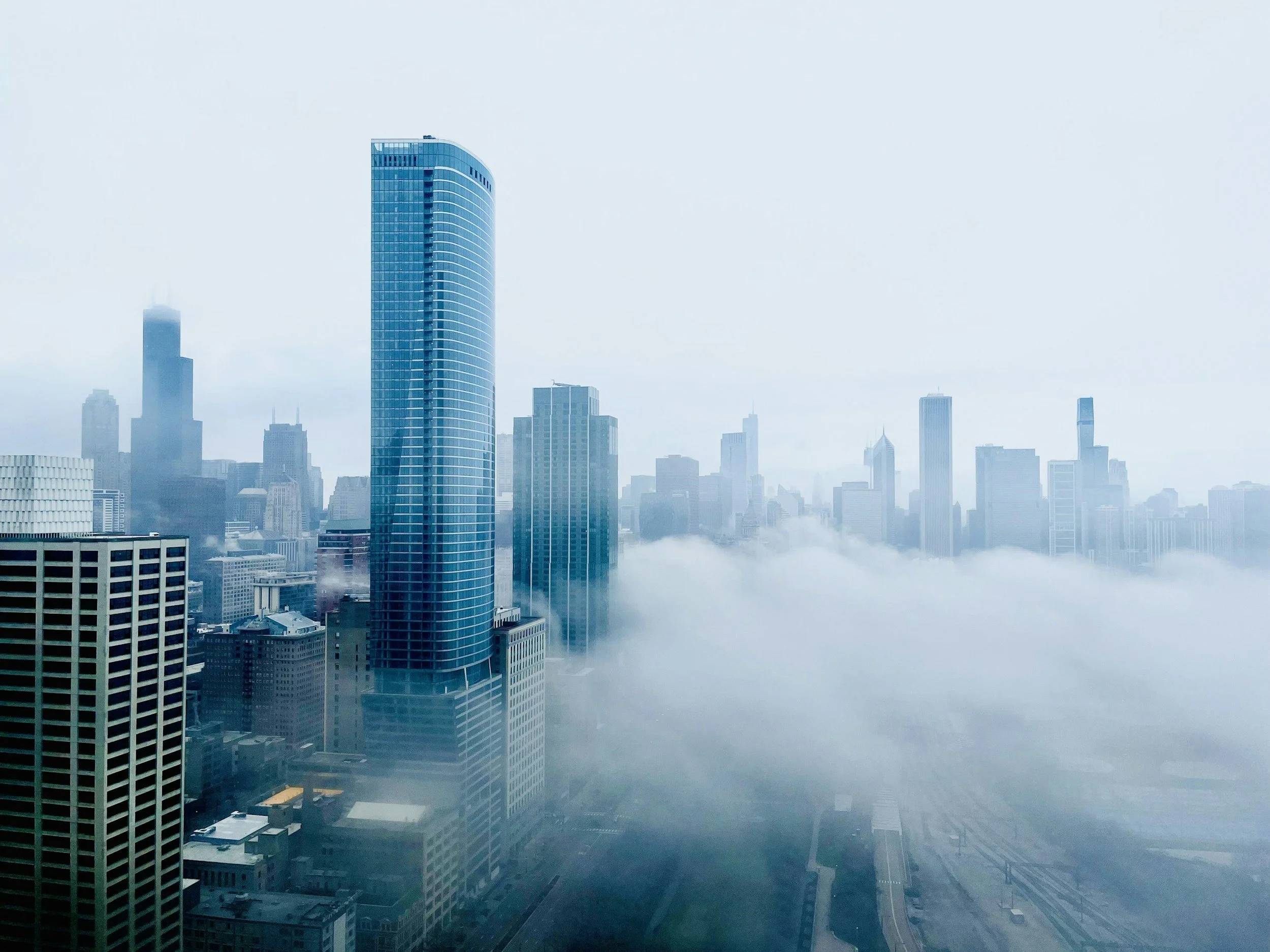 A city skyline with a tall modern blue glass skyscraper in the foreground, surrounded by other high-rise buildings partially obscured by fog or low clouds.