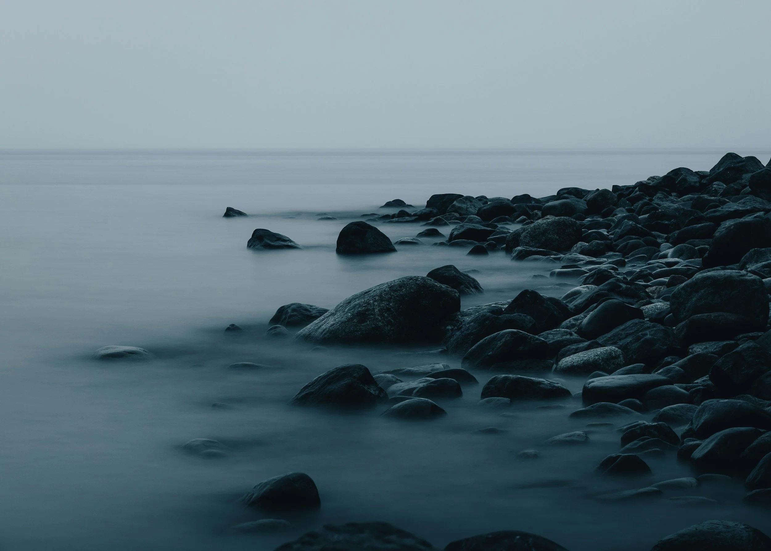 A long exposure photograph of a rocky shoreline with smoothed stones along the water's edge, under a foggy sky with calm, misty water.