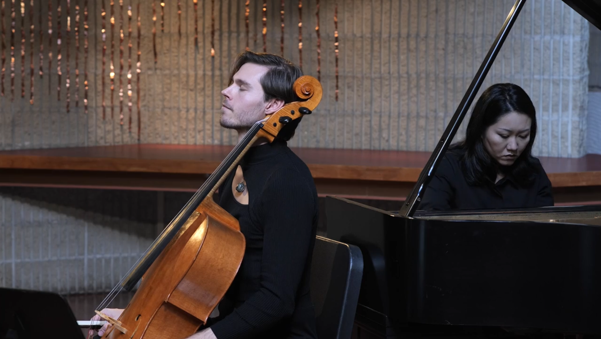 Nicholas Johnson playing the cello with his eyes closed, and Seiko Matsuzaki playing the piano with her head down, performing together in a music video.