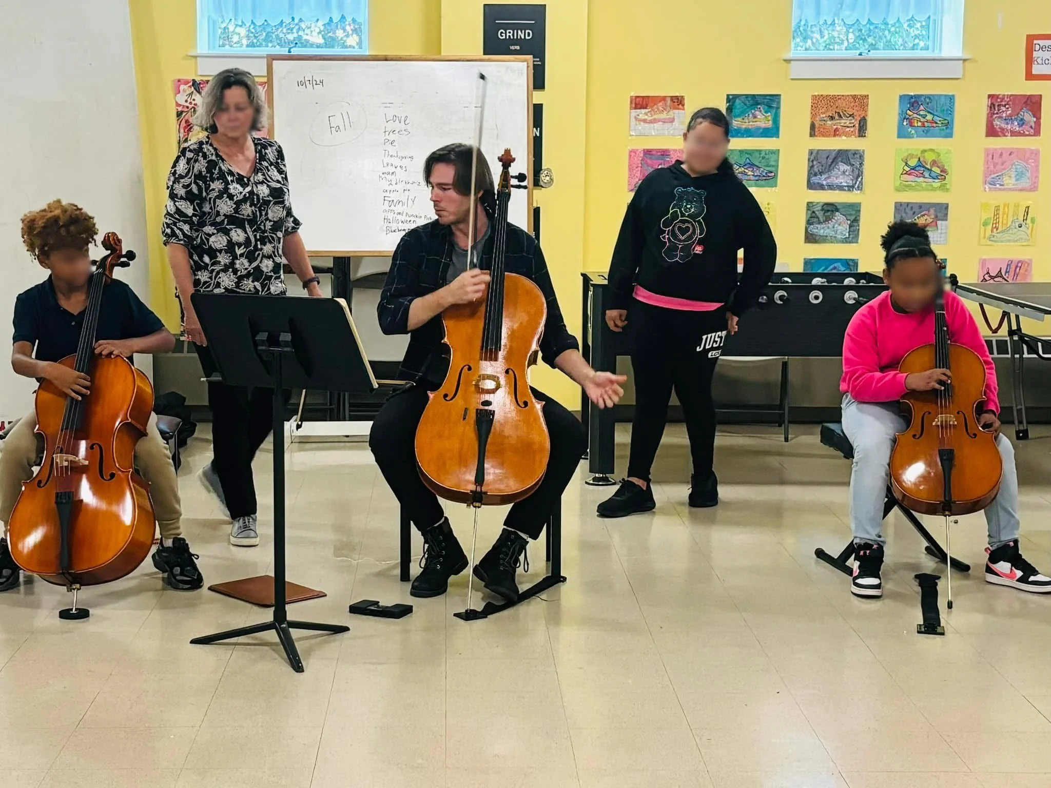 Group of five people, three children and two adults, in a room with colorful artwork on the walls. Cello instructor Nicholas Johnson is teaching a group cello class. The room has a whiteboard with handwritten notes about fall and family.