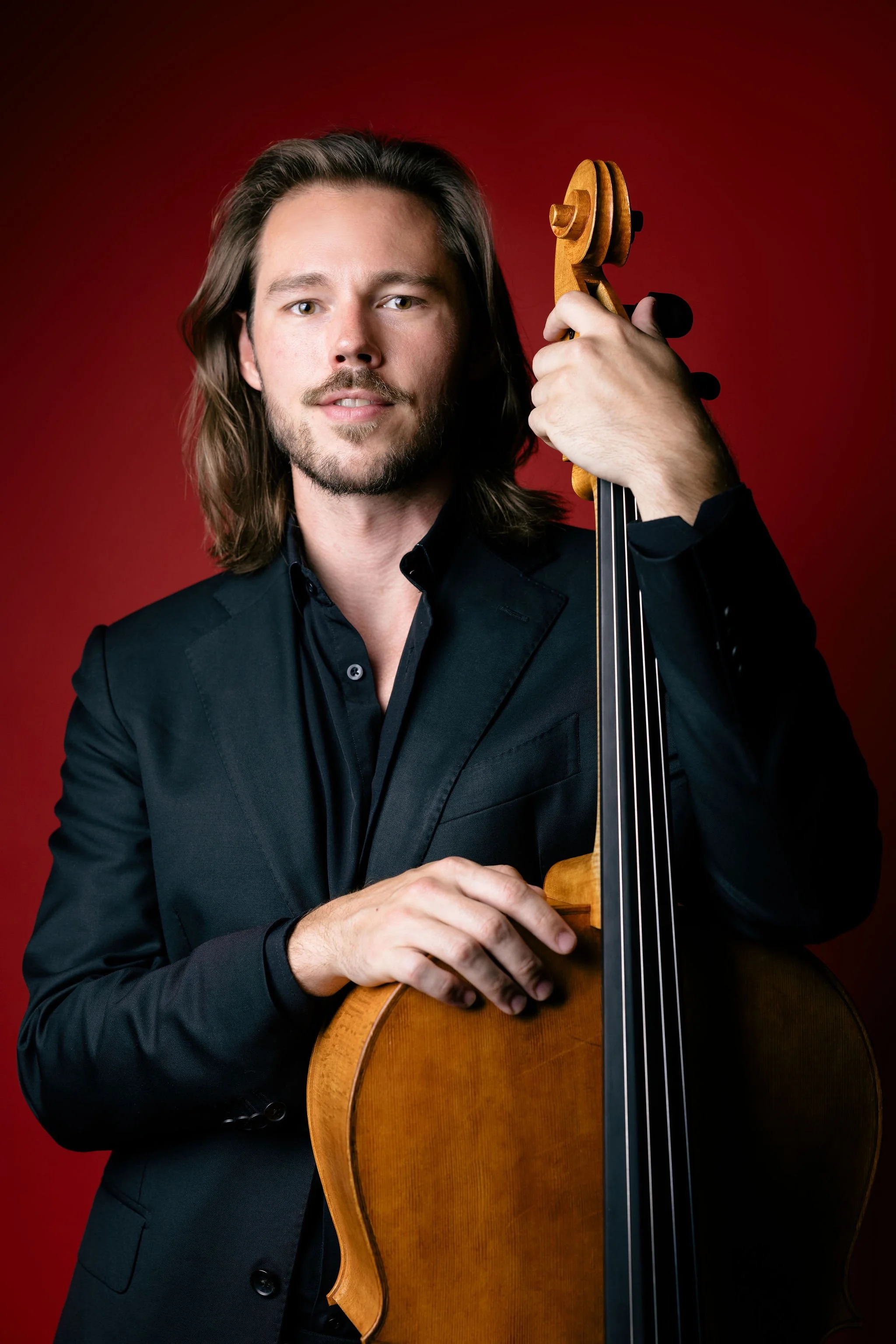 A man with long brown hair and a beard wearing a black suit jacket and shirt is holding a cello against a red background.