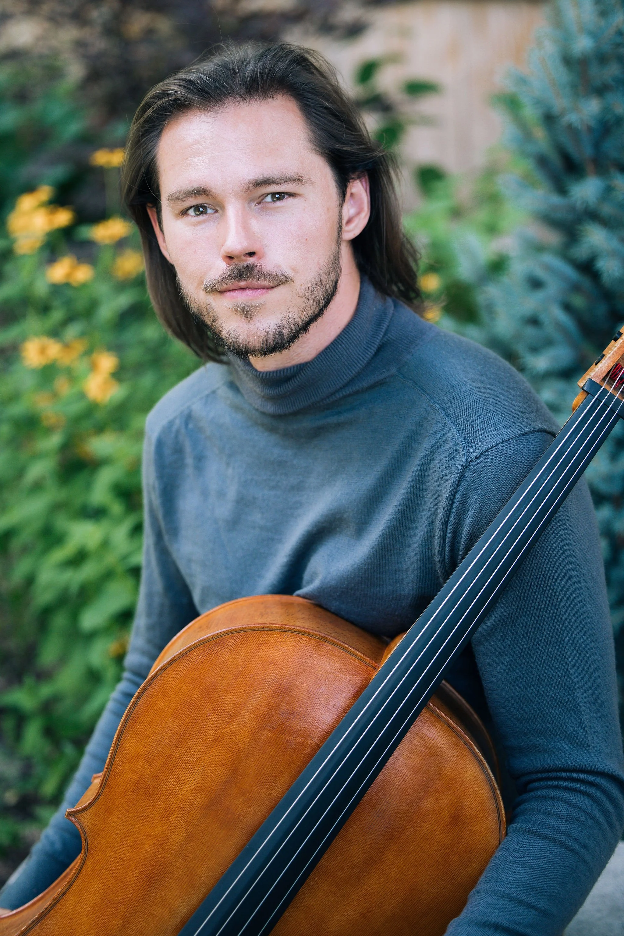 Nicholas Johnson with long brown hair and facial hair holding a classical cello outdoors with greenery and yellow flowers in the background.
