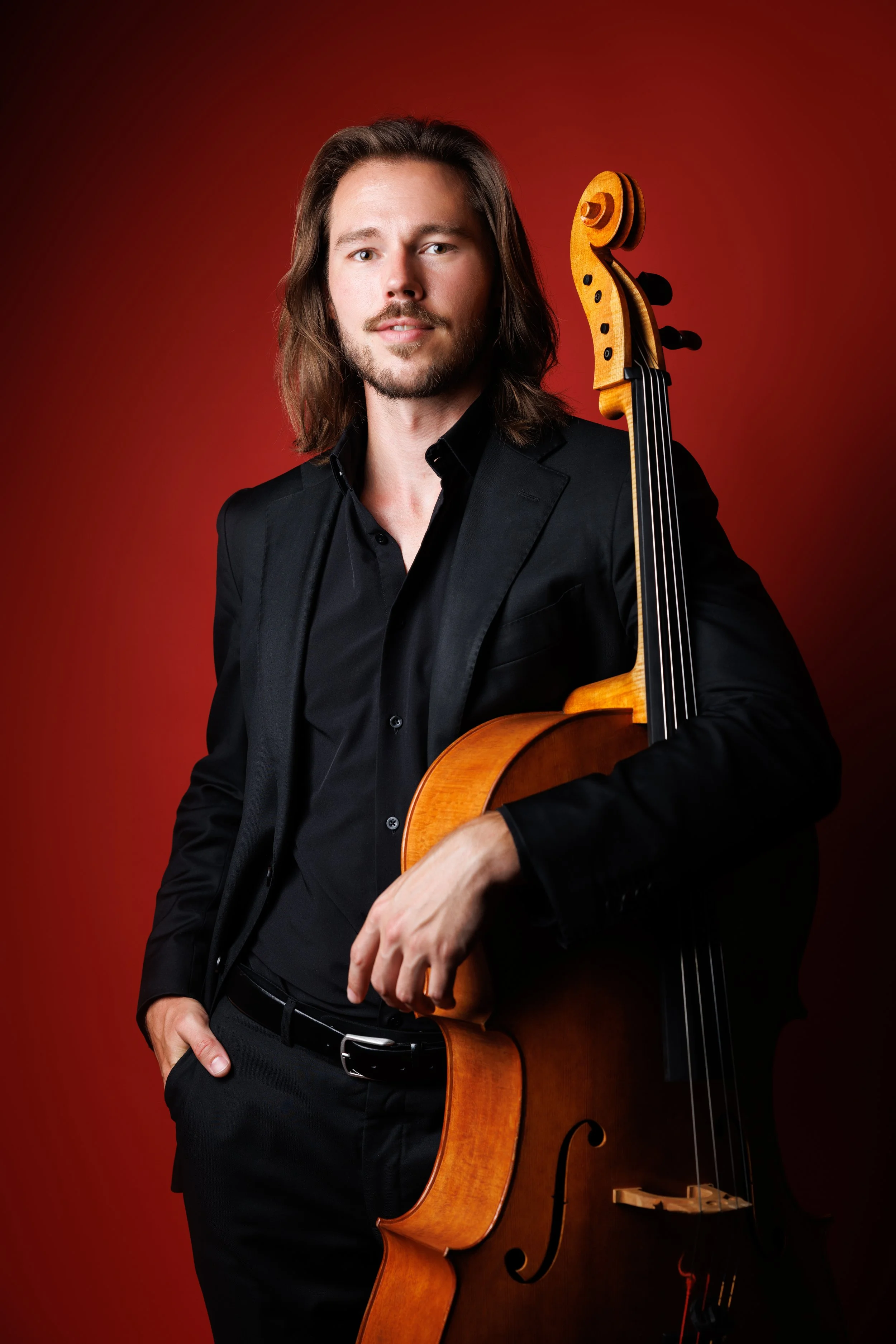Nicholas Johnson with cello, with long brown hair, dressed in a black suit against a red background.