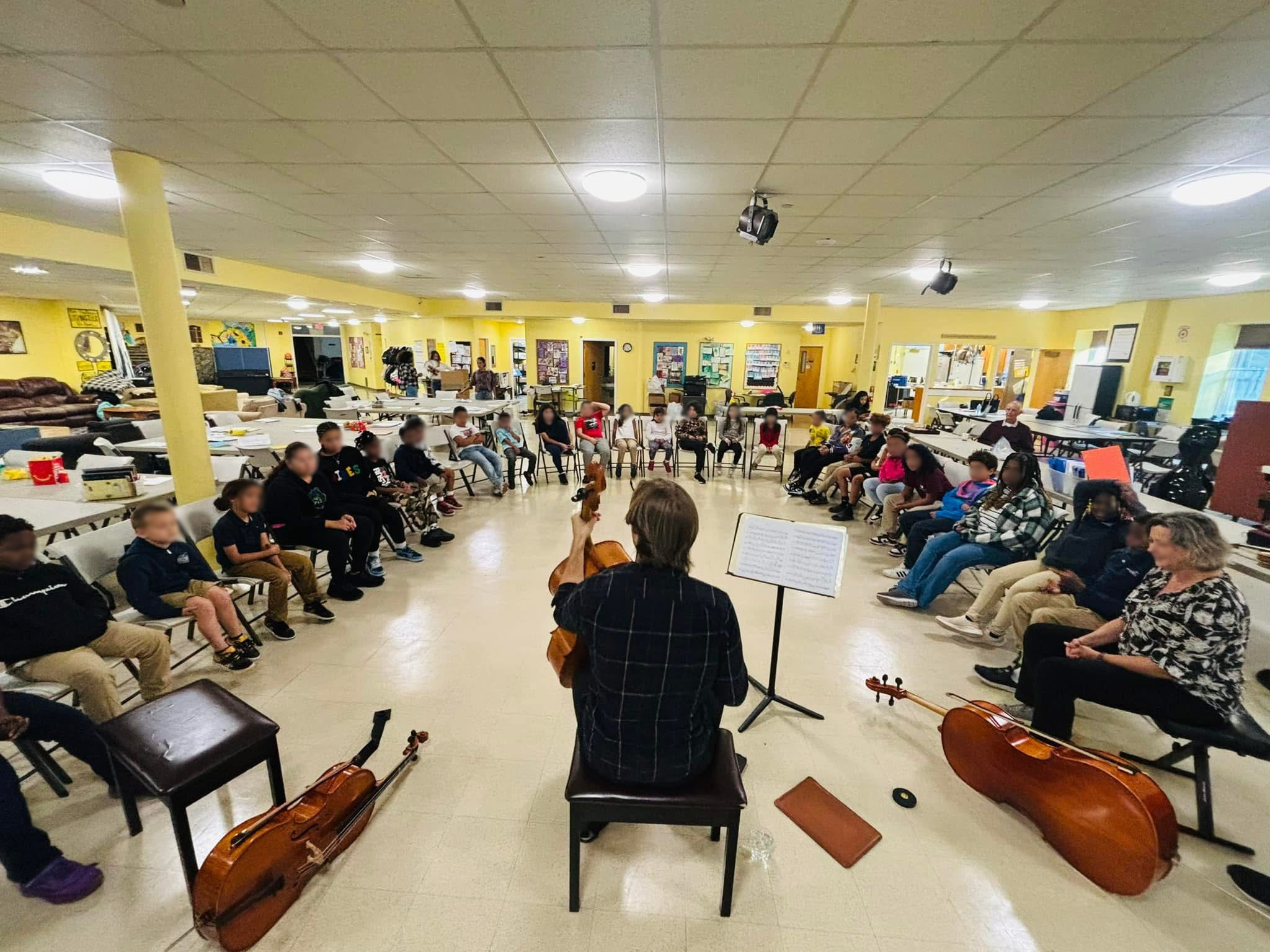 A group of children and adults sitting in a circle in a room, with a string instrument being played by Nicholas Johnson at the front.