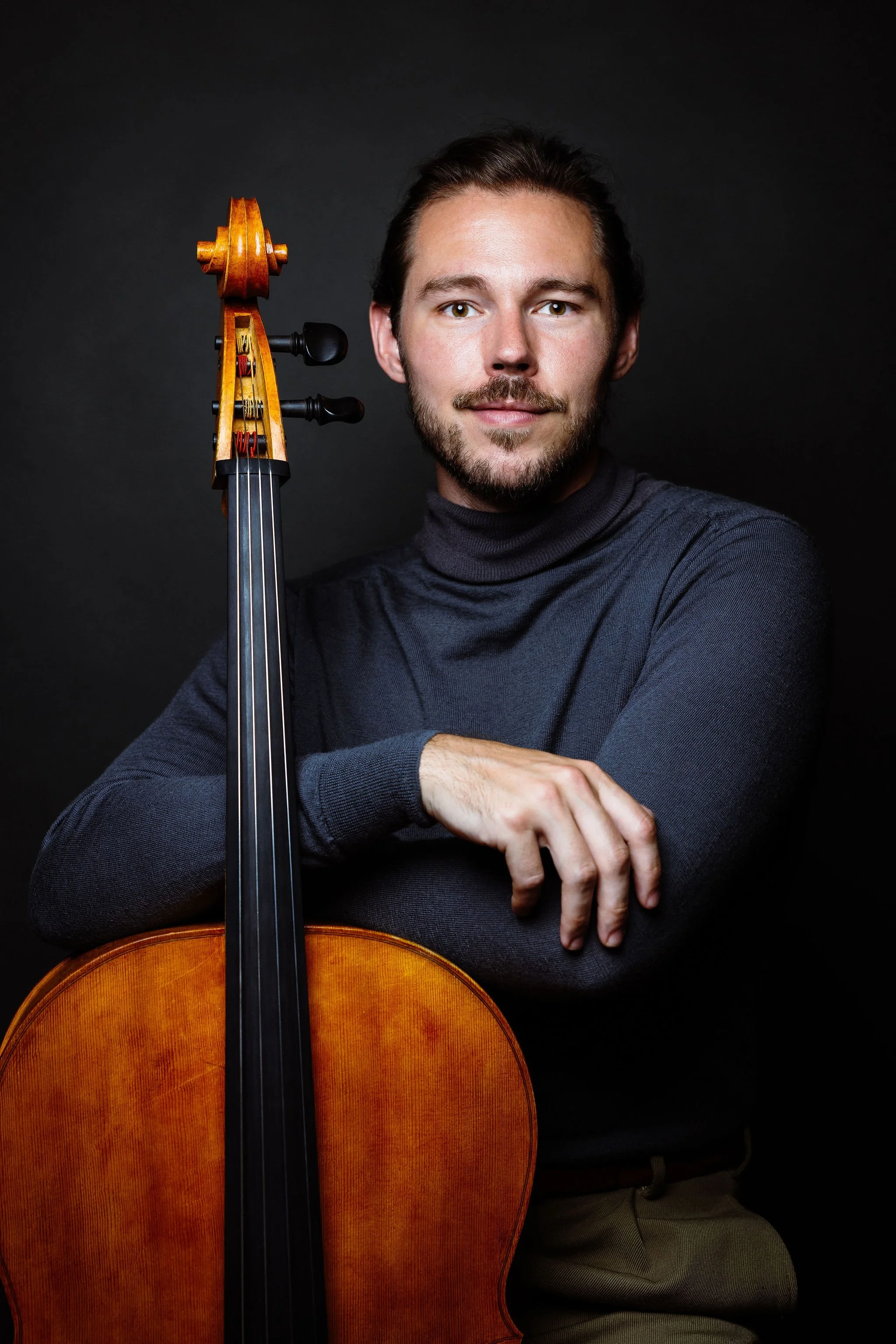 Nicholas Johnson Cello wearing a dark turtleneck, sitting next to a cello, with a dark background.