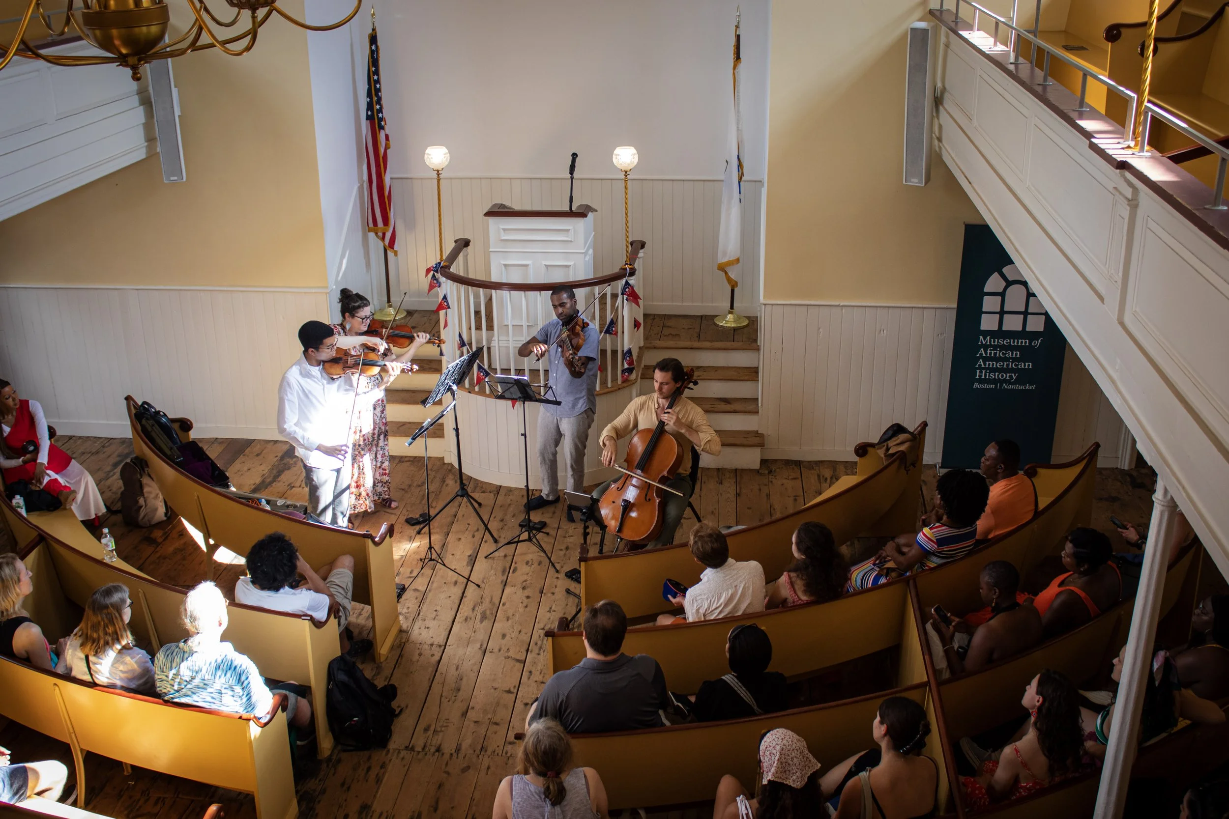 Nicholas Johnson, cellist, playing with Boston Public Quartet on a small stage at the Museum of African American History, with an audience seated in yellow chairs in a wood-floored room decorated with American flags.