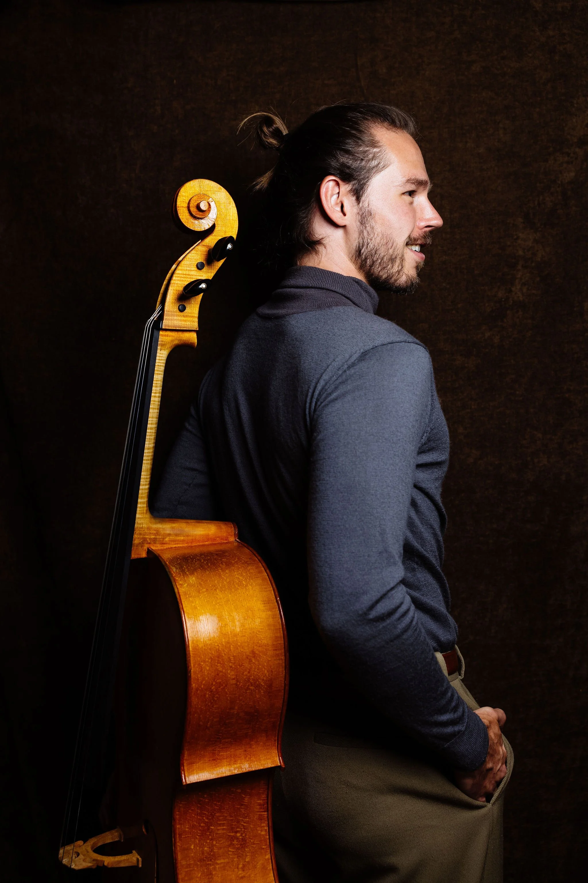 Nick Johnson, Cellist, with a beard and long hair tied in a bun, wearing a dark gray shirt, is standing with a wooden cello resting on his back. He is smiling and looking to his right.