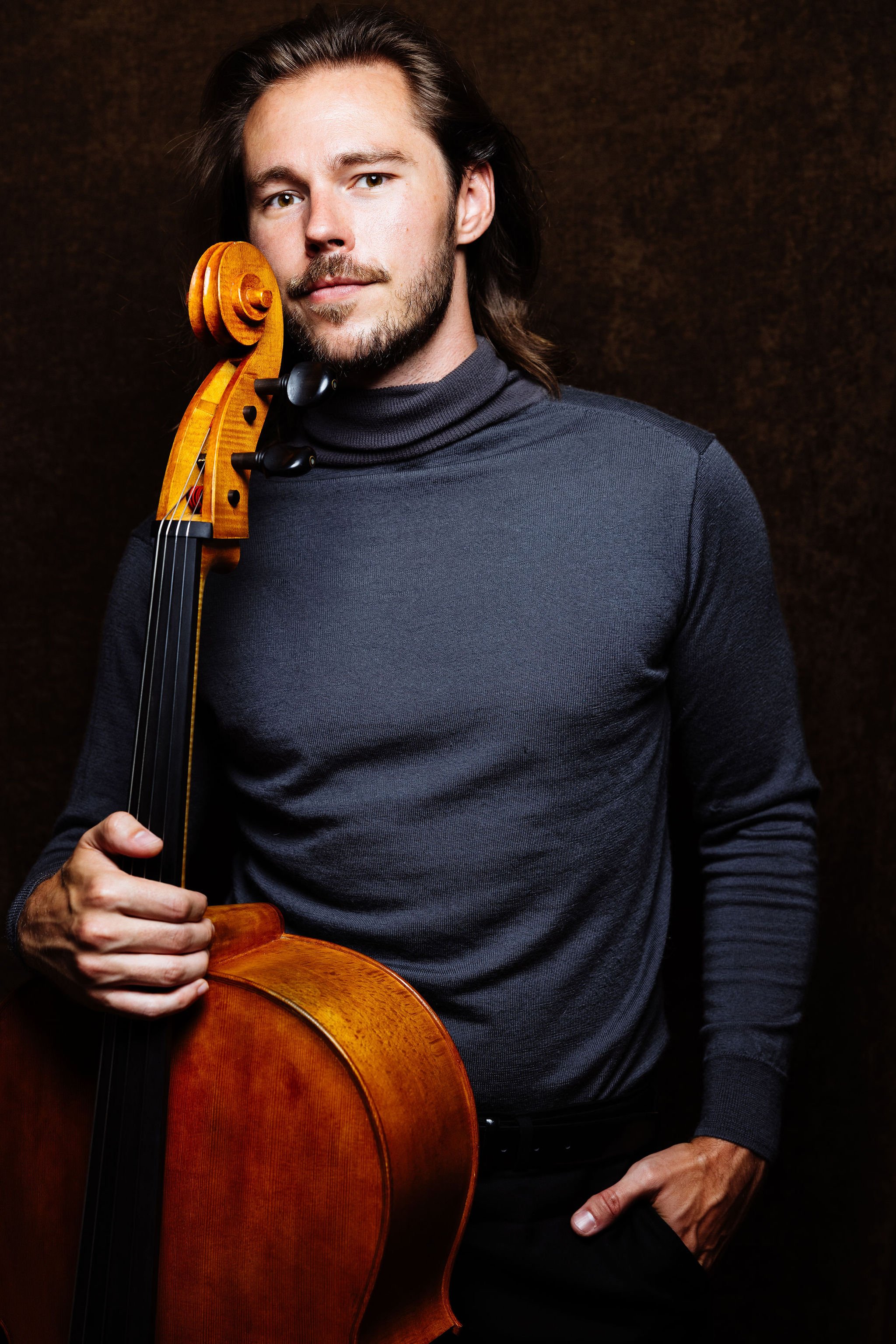 Nicholas Johnson, Cellist, with long brown hair and beard holding a wooden cello against a dark background.