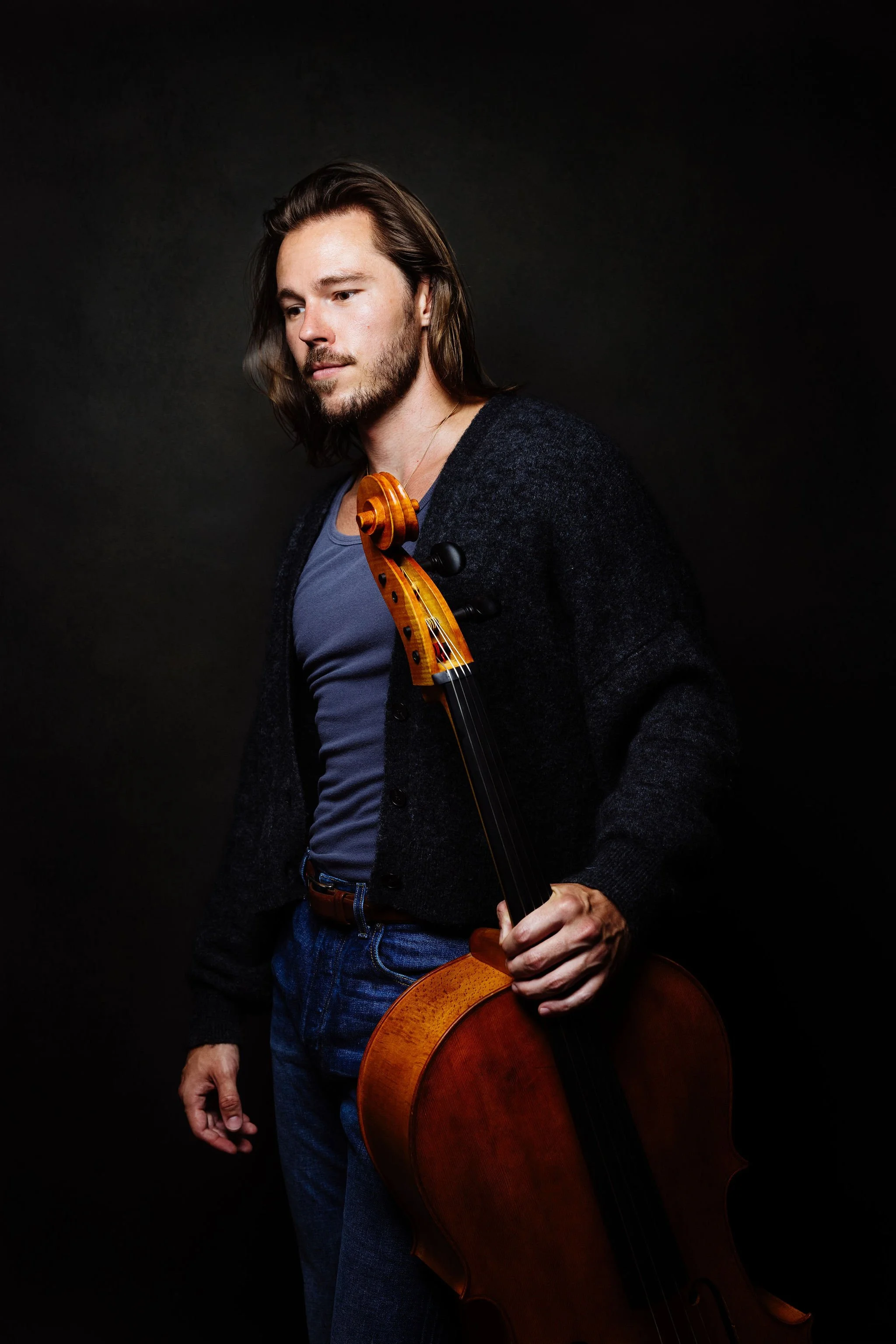 Nicholas Johnson dressed in a dark sweater and blue jeans, holding a cello against a dark background.