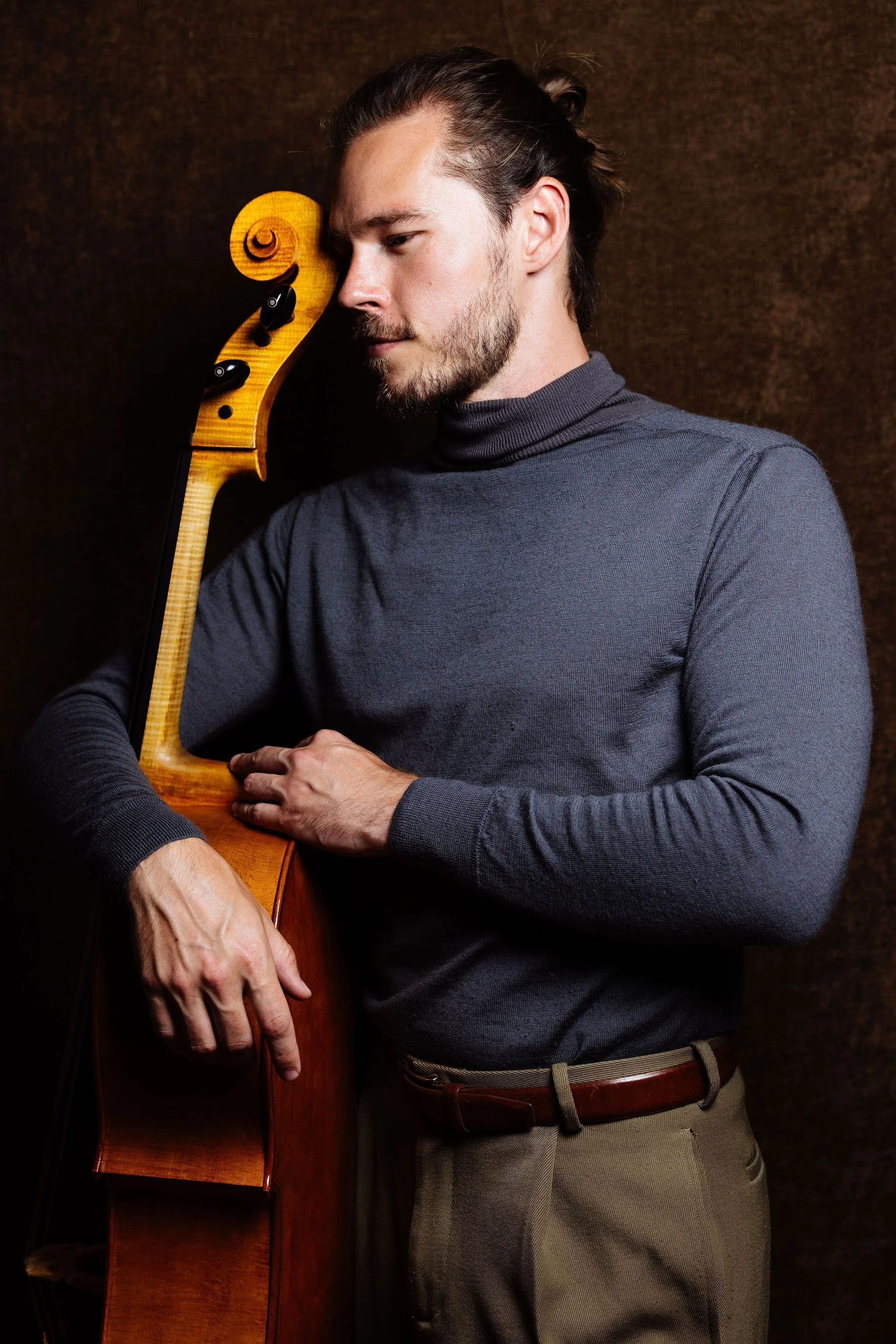 Cellist Nicholas Johnson with a beard and long hair tied back, wearing a gray turtleneck and green pants with a brown belt, is holding a cello close to his face, with the scroll resting against his forehead, against a dark brown background.