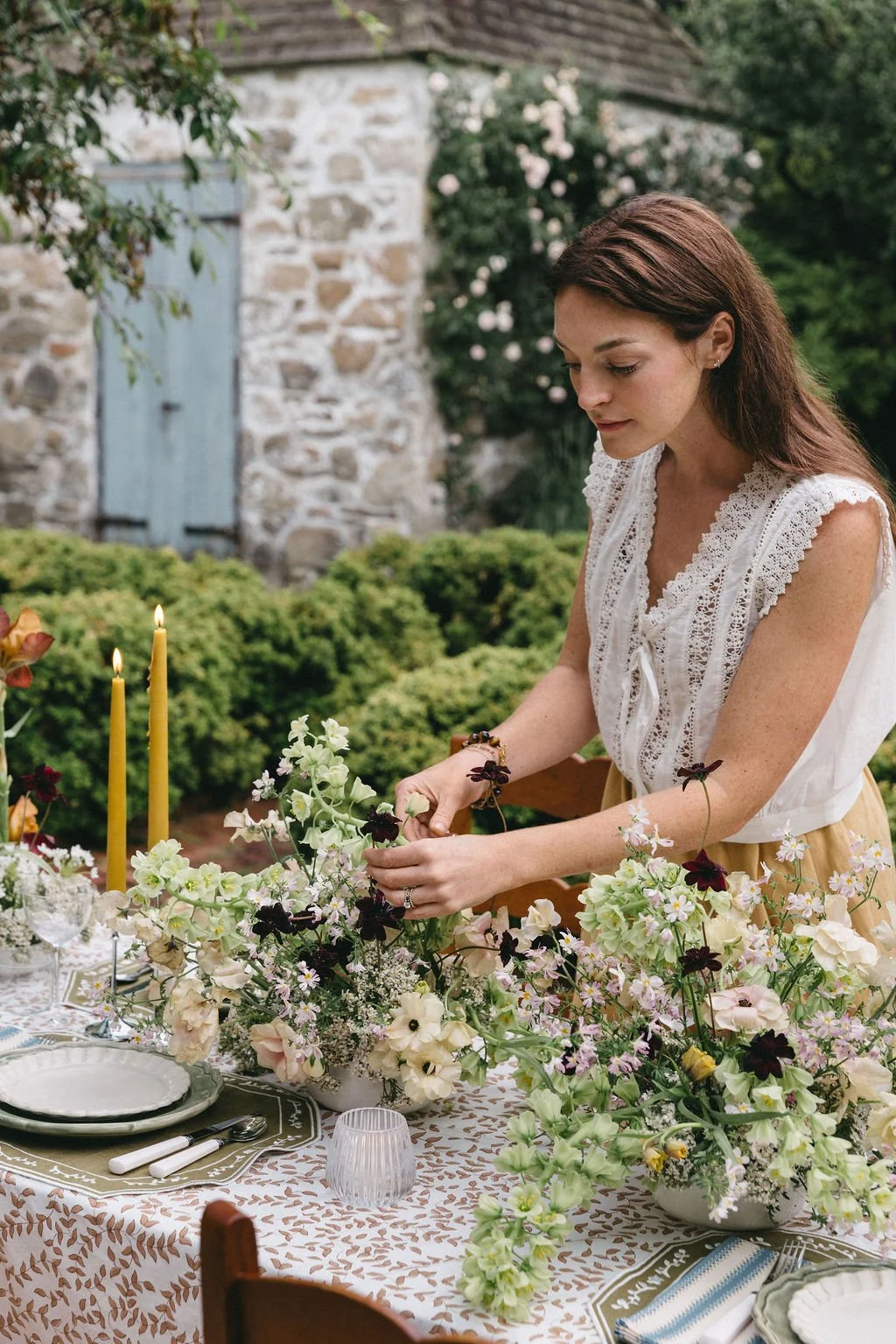 A woman arranging flowers on an outdoor table decorated with floral arrangements, candles, and tableware, with a stone building and greenery in the background.