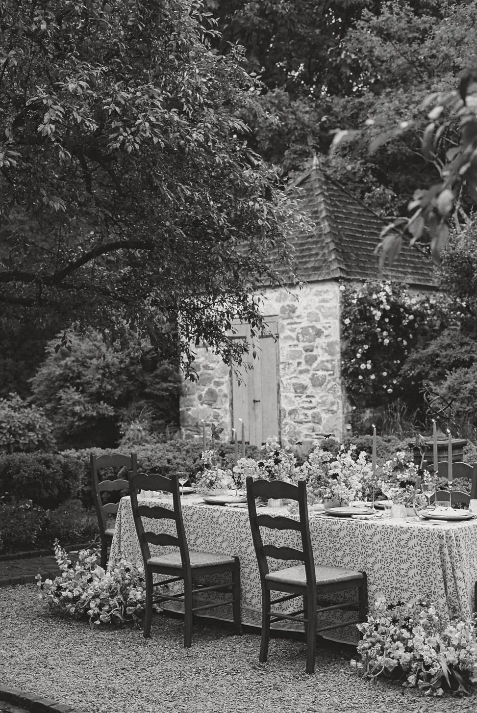 An outdoor garden setting with a long table decorated with a patterned tablecloth, floral arrangements, and candles. Wooden chairs surround the table, which is set for a meal. In the background, there is a small stone building with a wooden door and a tiled roof, surrounded by trees and foliage.