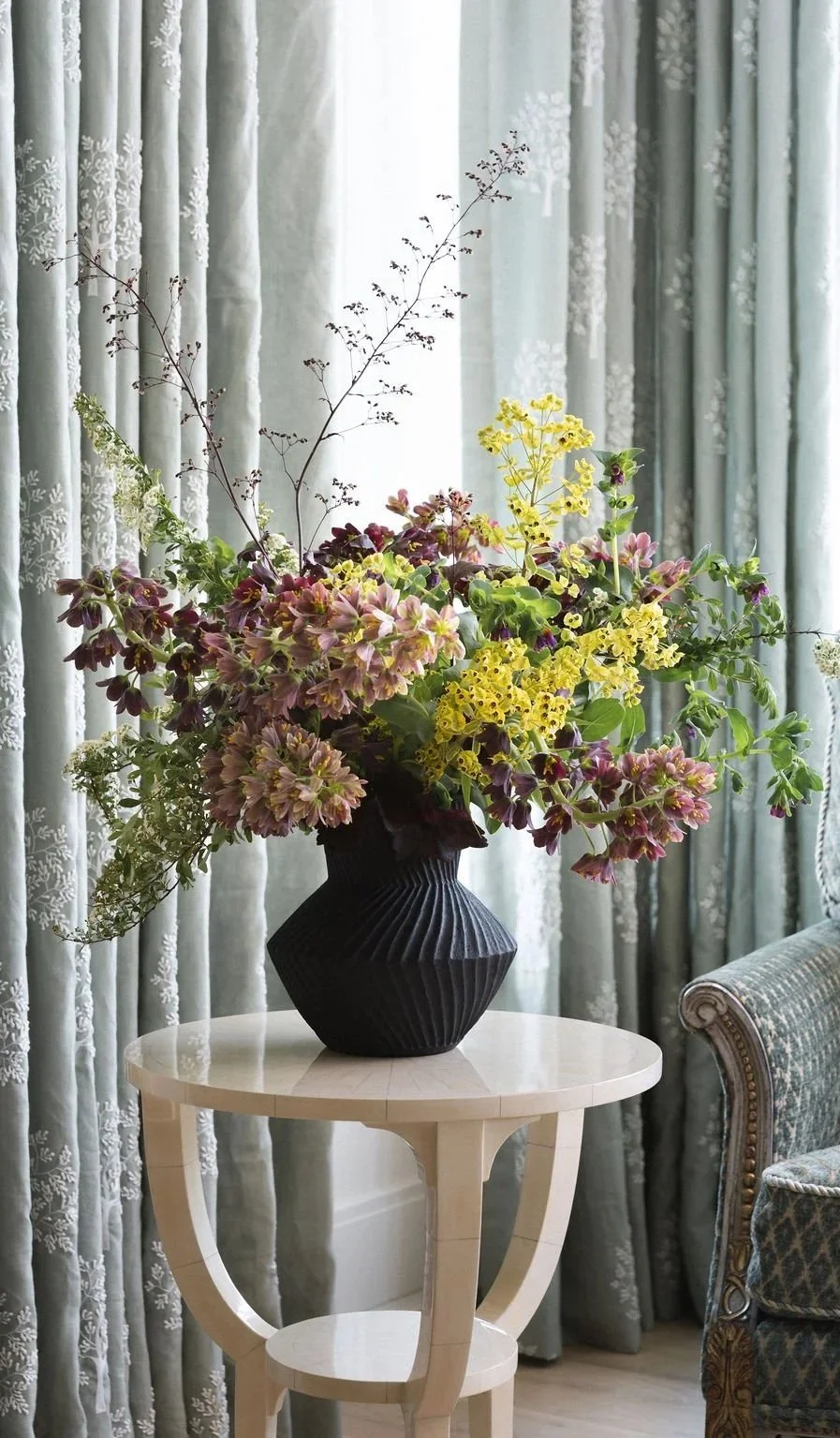 A black textured vase with a large bouquet of pink, purple, and yellow flowers sits on a small white side table, with patterned curtains and a patterned upholstered chair in the background.