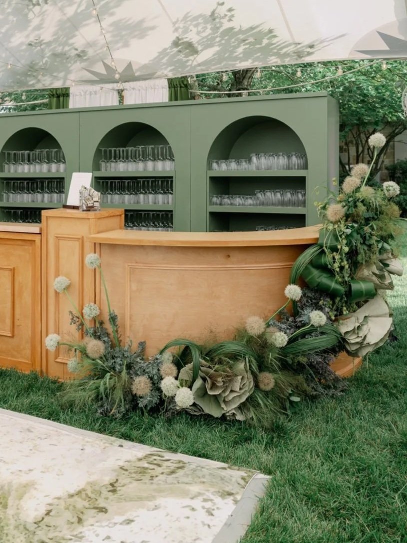 A wooden bar counter with green shelving behind it at an outdoor event. The shelves hold clear glasses, and a floral arrangement with large leaves and soft round white flowers decorates the front of the bar.