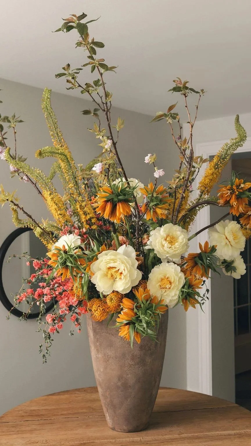 A large ceramic vase filled with a mixed floral arrangement including white peonies, orange lilies, pink blossoms, and tall yellow and green flowers on a wooden table.