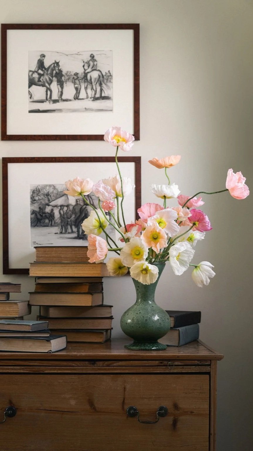 A green vase with pink, white, and peach poppy flowers on a wooden table with a stack of books. Two black-and-white sketch drawings of horses, one above the other, are hung on the beige wall behind the table.