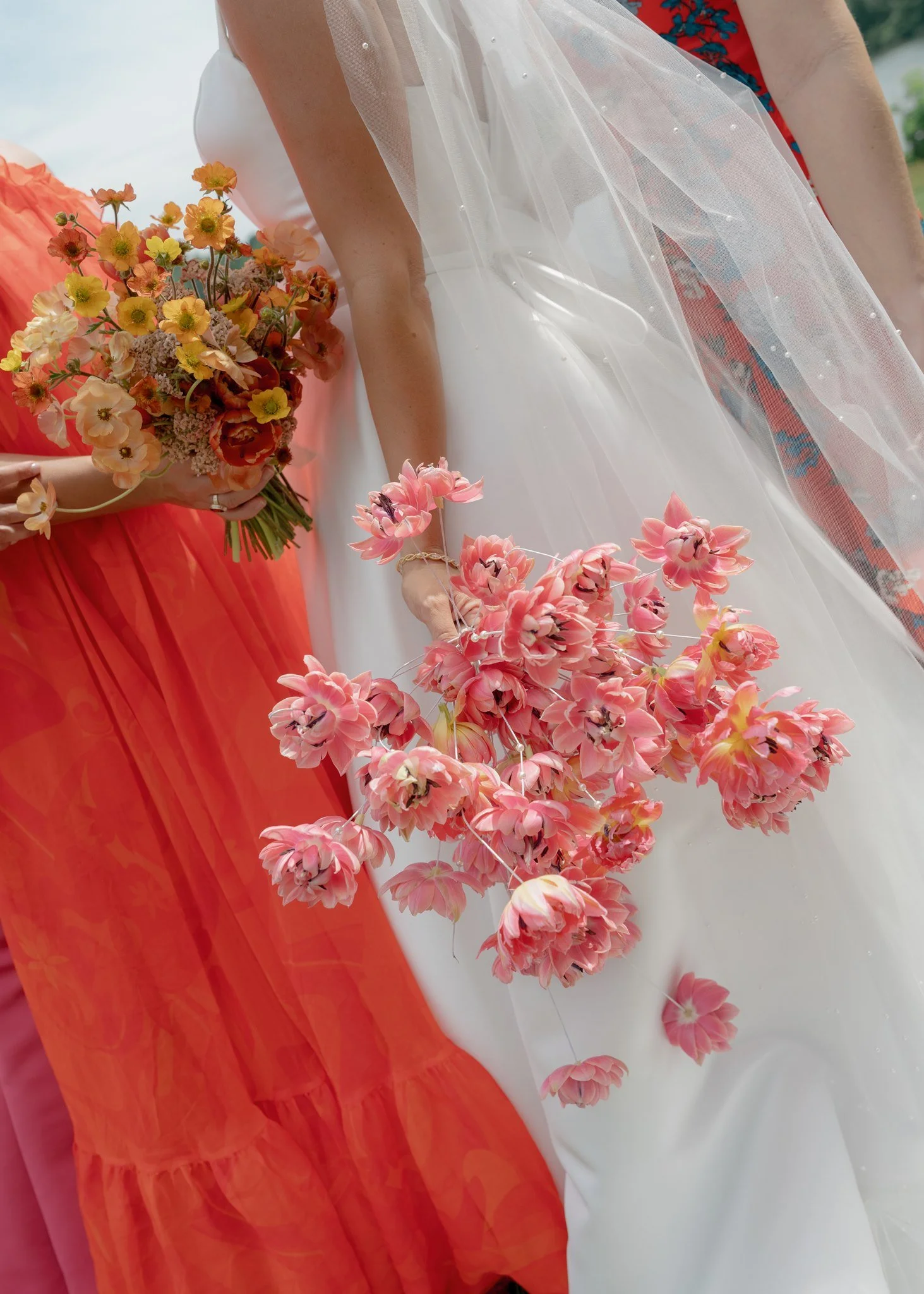 Close-up of people holding pink and peach flowers at a wedding or formal event, with a woman in a white dress and veil and another person in an orange dress holding a bouquet of flowers.