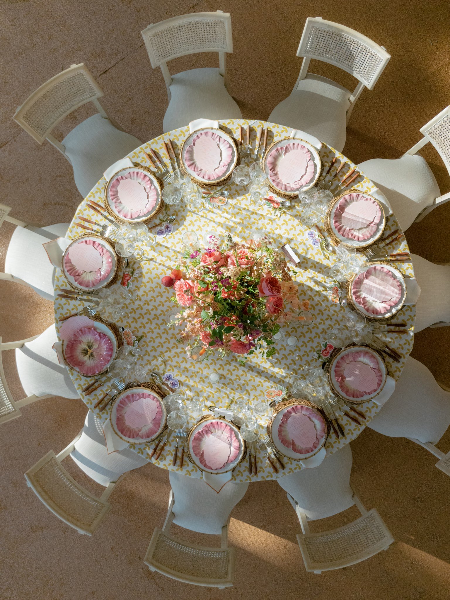 Round dining table set for a meal with a yellow patterned tablecloth, pink and gold plates with ruffled edges, multiple wine glasses, gold cutlery, and a floral centerpiece with pink and peach flowers.