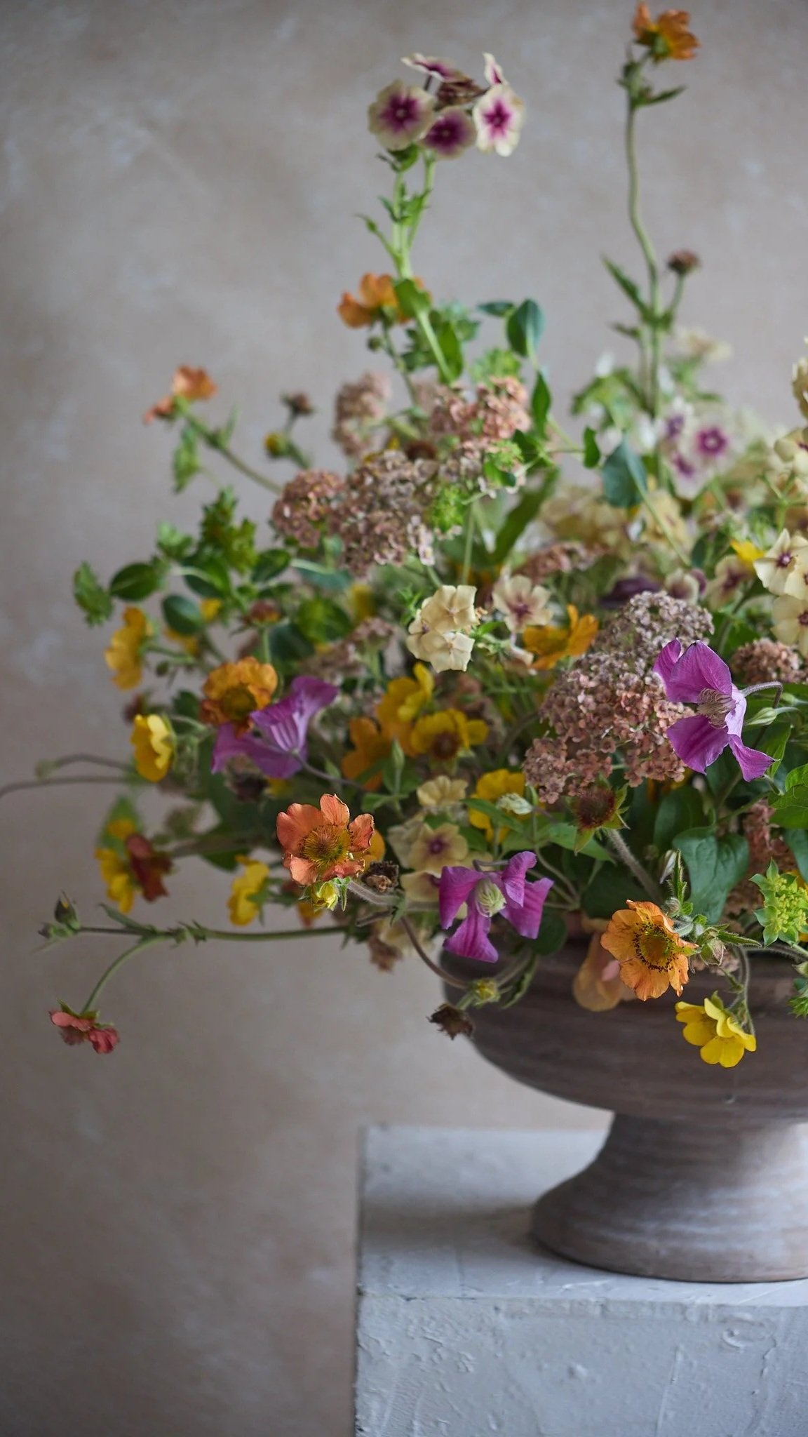 A large, rustic bowl filled with colorful real flowers including pink, yellow, purple, and orange, set on a white stand against a beige wall.