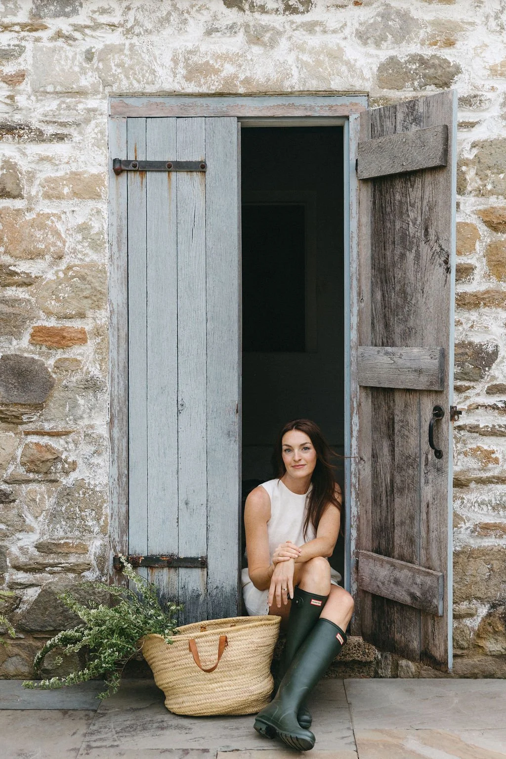 A woman sitting in the doorway of a rustic stone building with wooden shutters, wearing a sleeveless white top, shorts, and green rain boots, with a woven basket and greenery beside her.