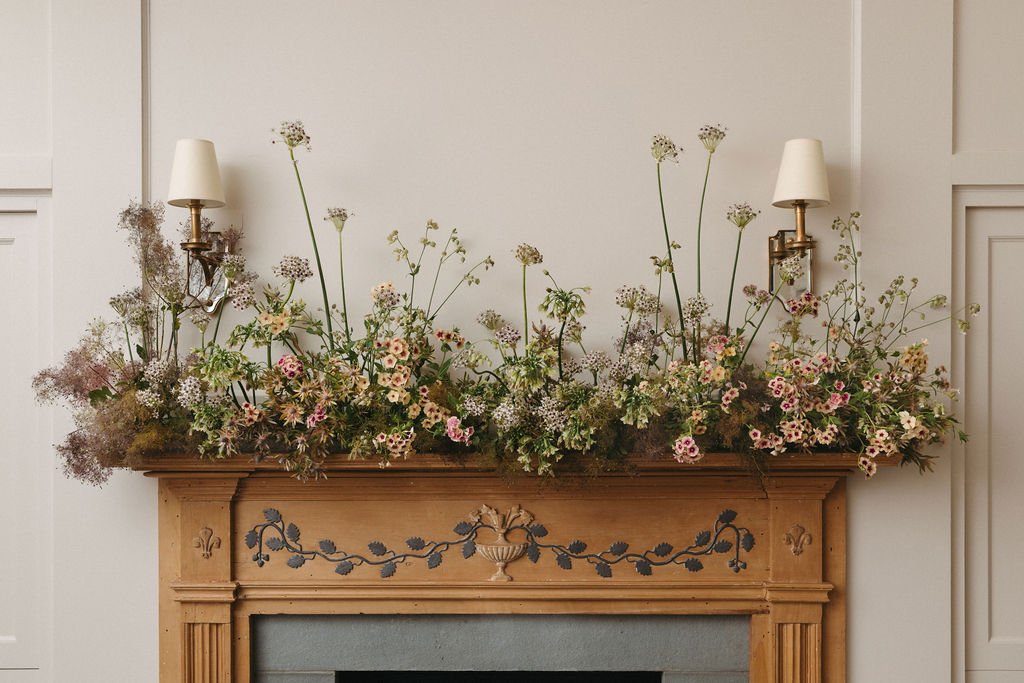 Decorative fireplace mantel with a large arrangement of wildflowers and greenery, flanked by wall-mounted lamps with white shades.