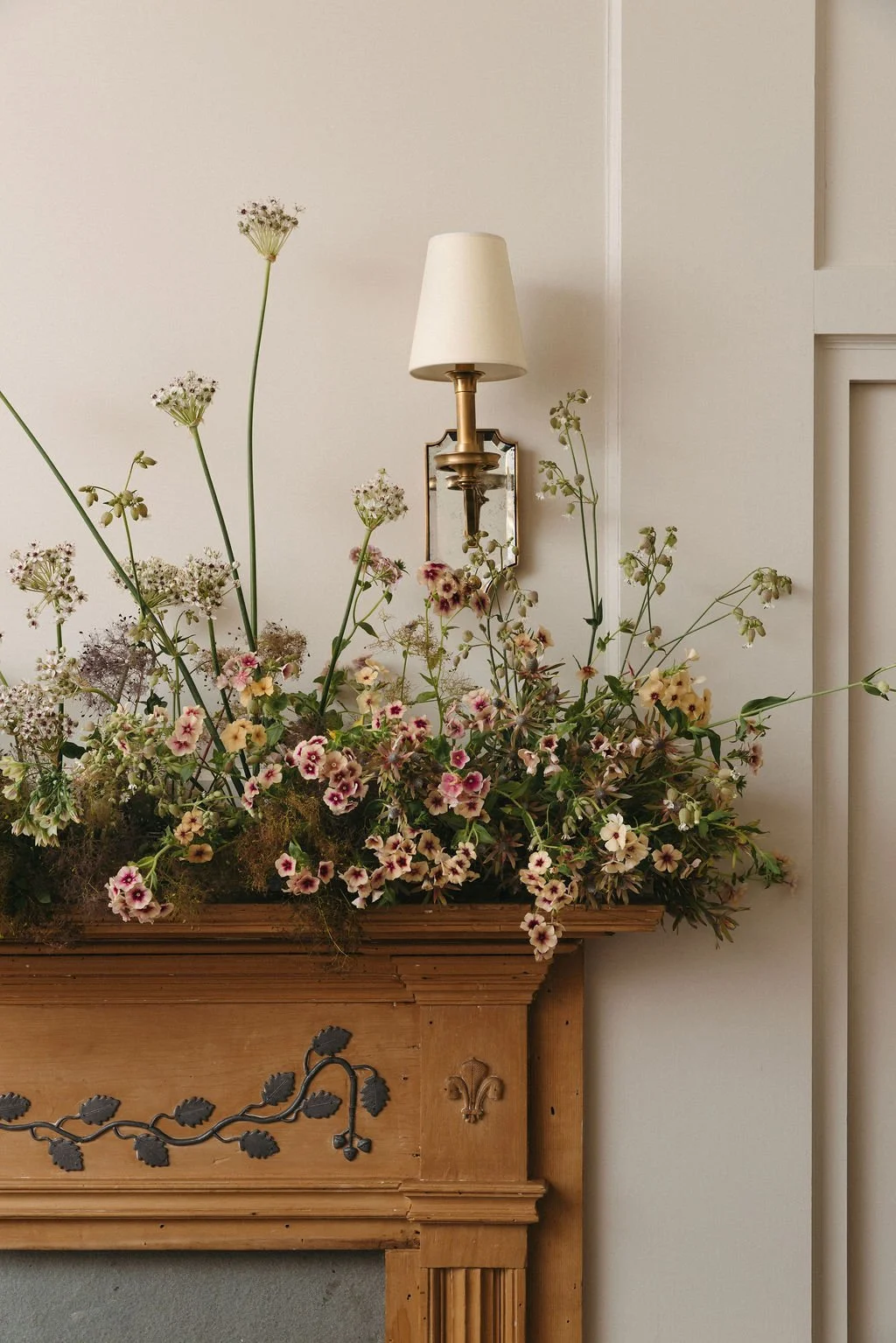 A floral arrangement with various green-stemmed flowers and small pink and white blossoms on a wooden fireplace mantel, with a wall-mounted brass and white lampshade sconce above.