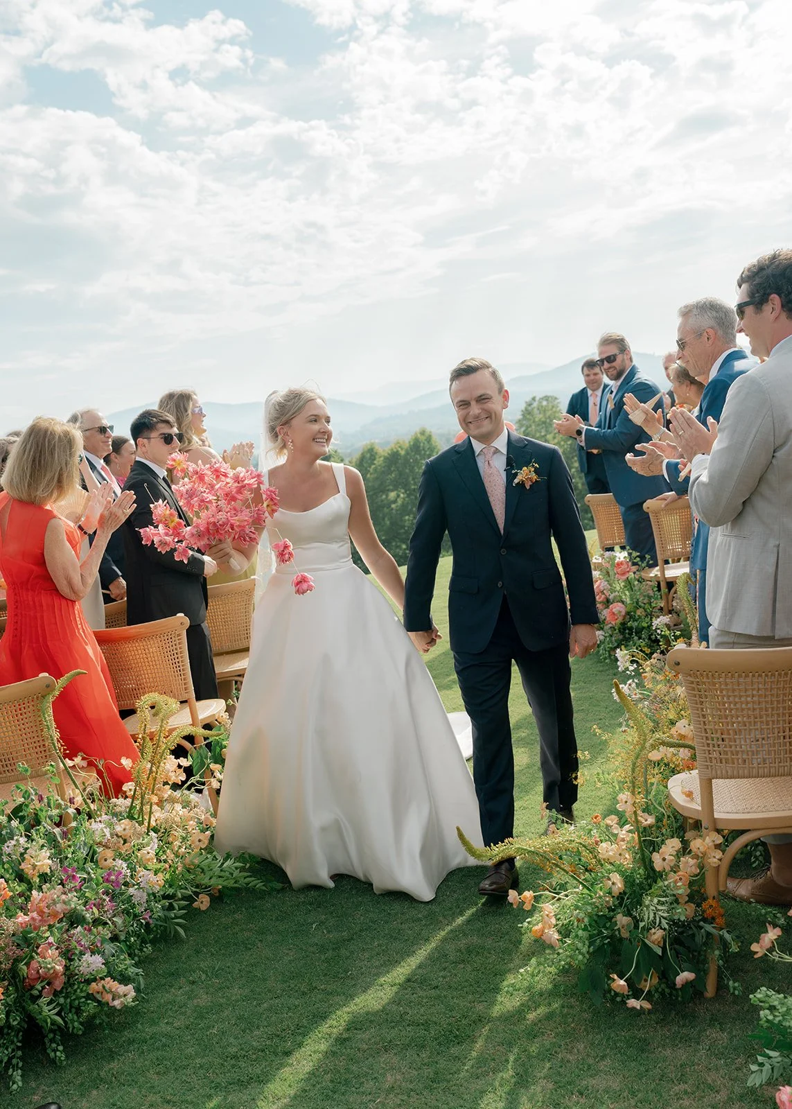 A bride and groom walk hand-in-hand down the aisle at an outdoor wedding ceremony. The wedding guests are clapping and smiling, with beautiful landscape and mountains in the background.
