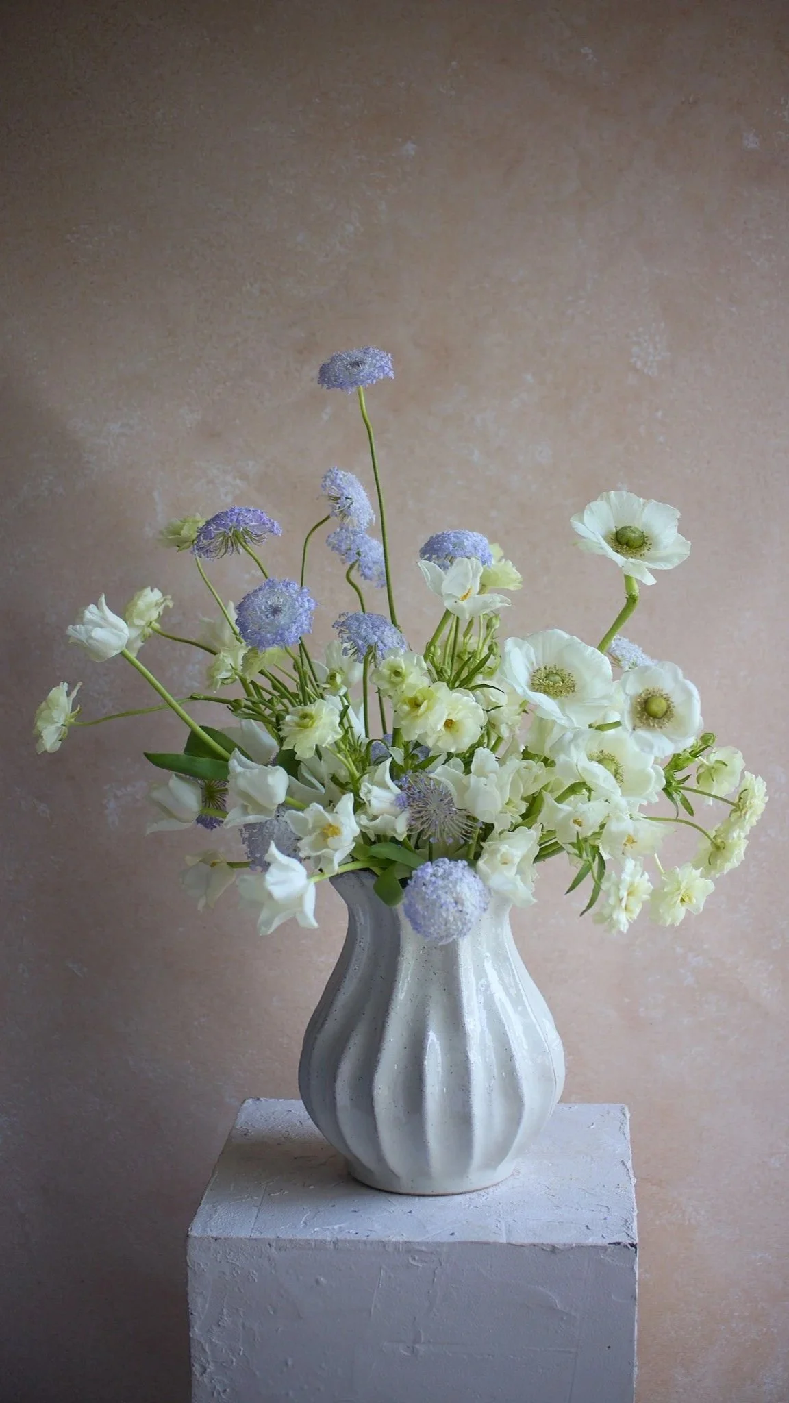A white vase with vertical ridges holds a bouquet of white and purple flowers. The vase is placed on a white pedestal against a beige wall with a textured surface.