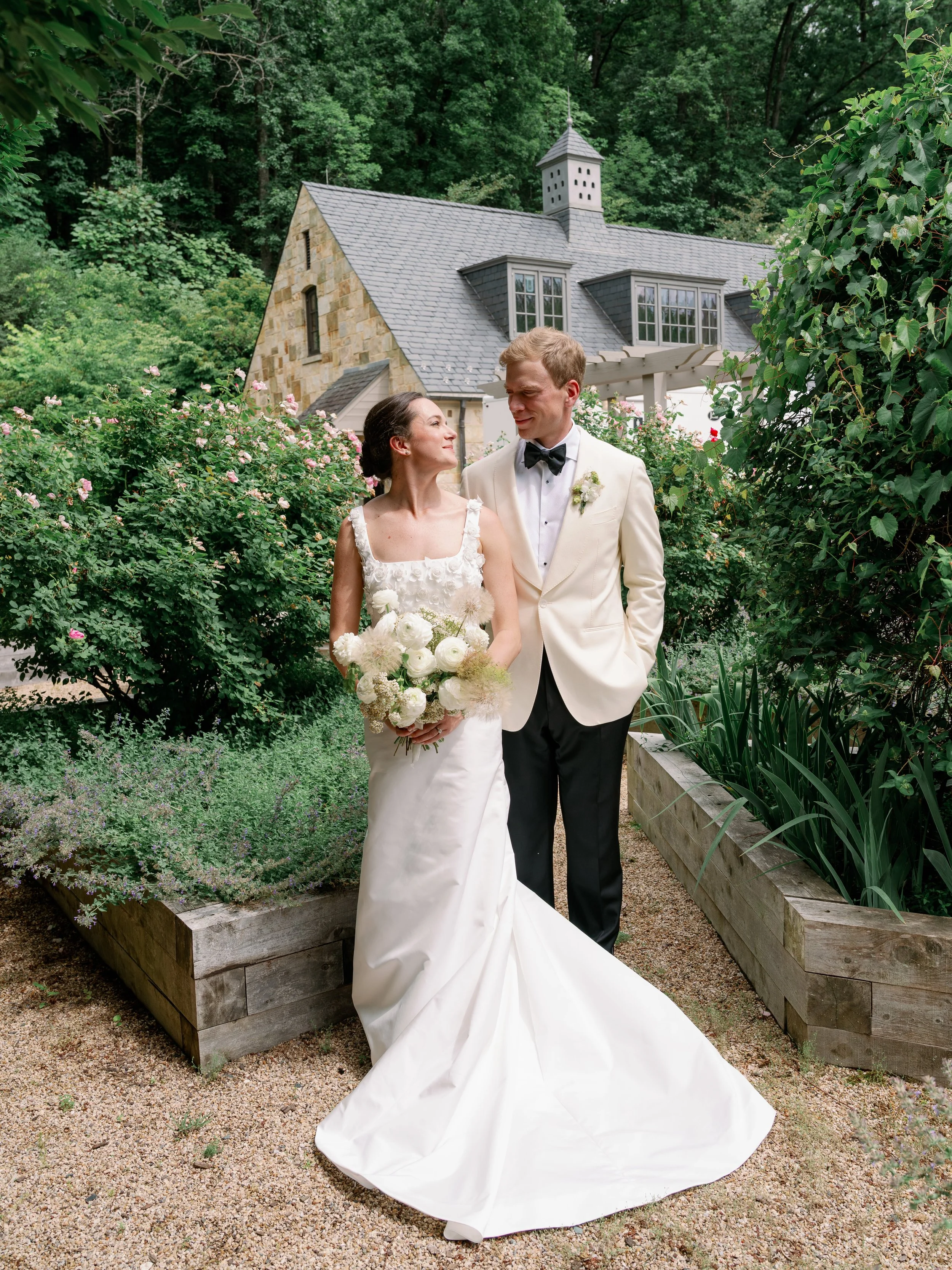 A bride in a white wedding gown holding a bouquet standing beside a groom in a white tuxedo with black bowtie, in a garden with a stone and wood house and lush green trees in the background.