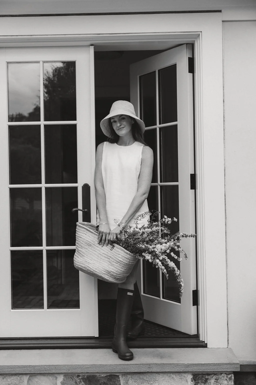 A woman standing in front of open double doors, holding a large woven basket with flowers inside, wearing a sleeveless dress, wide-brimmed hat, and rain boots in black and white.