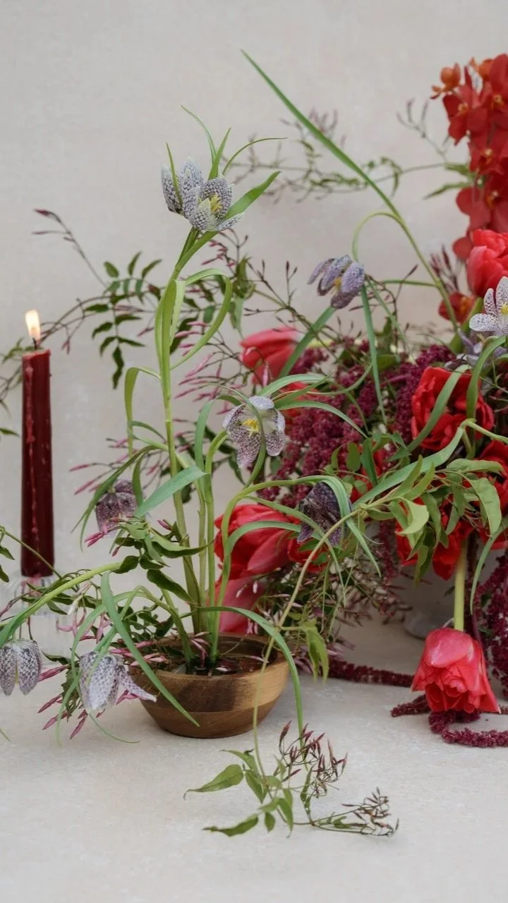 A small wooden bowl containing a flower arrangement with tall green grass and delicate purple flowers, surrounded by other pink and red flowers, a red candle, and a cream-colored background.