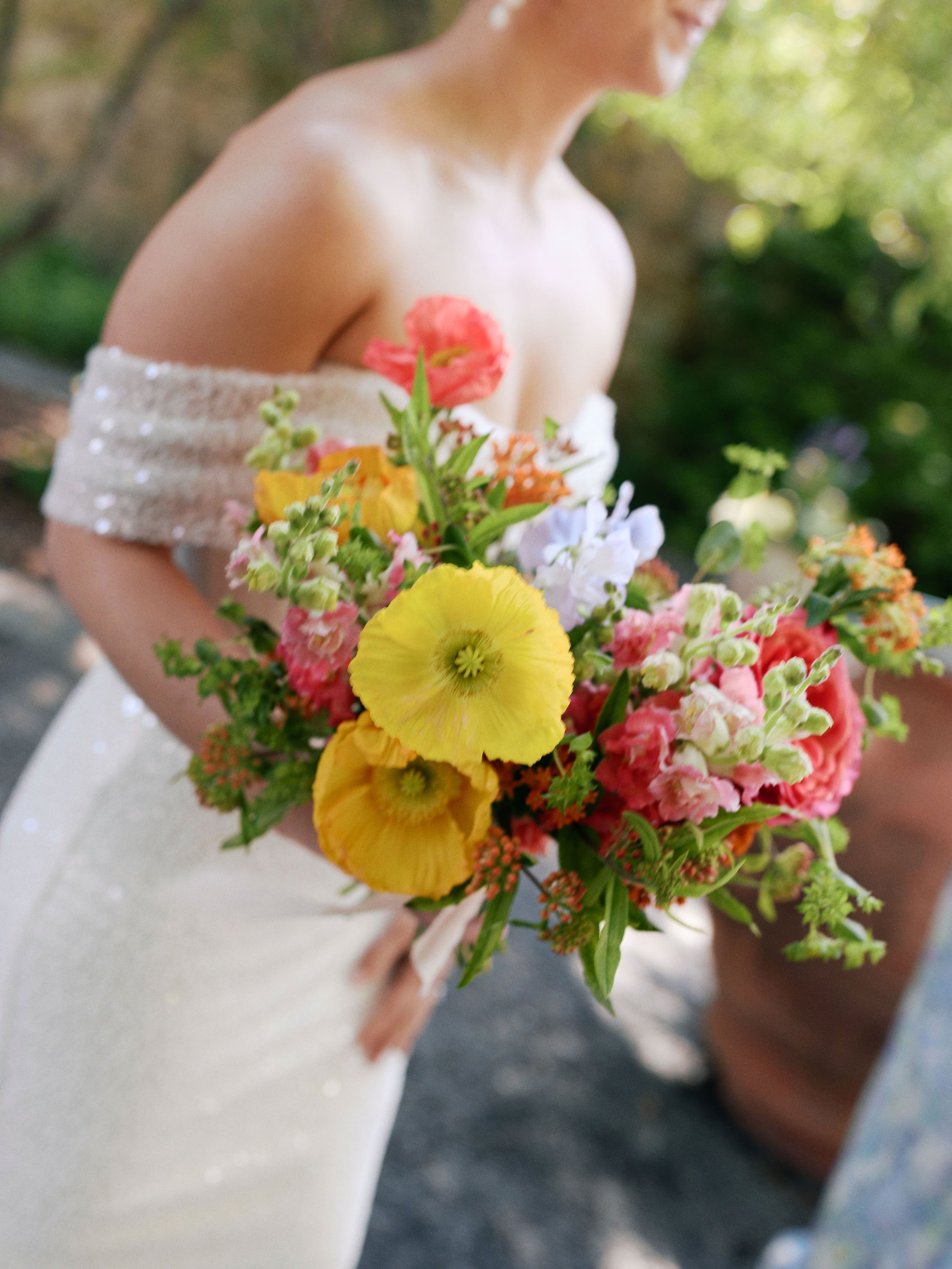 A woman wearing a white dress with small beads, holding a colorful bouquet of flowers with pink, yellow, and white blossoms, outdoors with a blurred background of greenery.