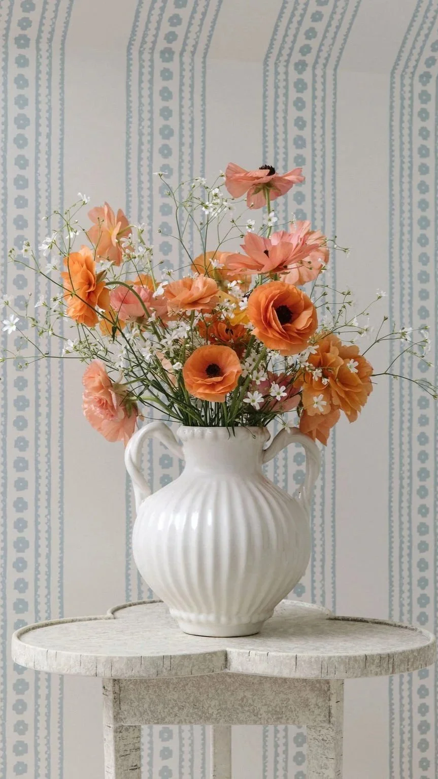 A white ceramic vase with vertical ridges and two handles, filled with pink and orange flowers, sits on a round, distressed white table against a light blue patterned wallpaper background.