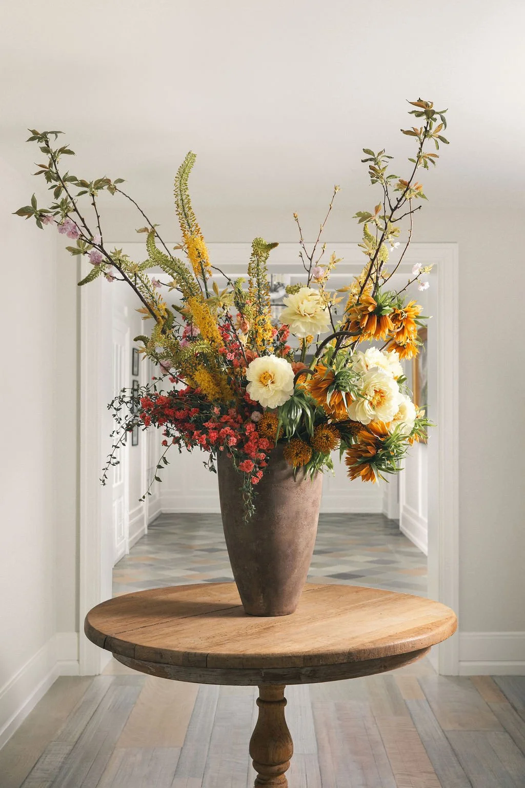 A tall floral arrangement in a large, rustic vase on a round wooden table in a bright, minimalistic hallway with white walls and a patterned floor.