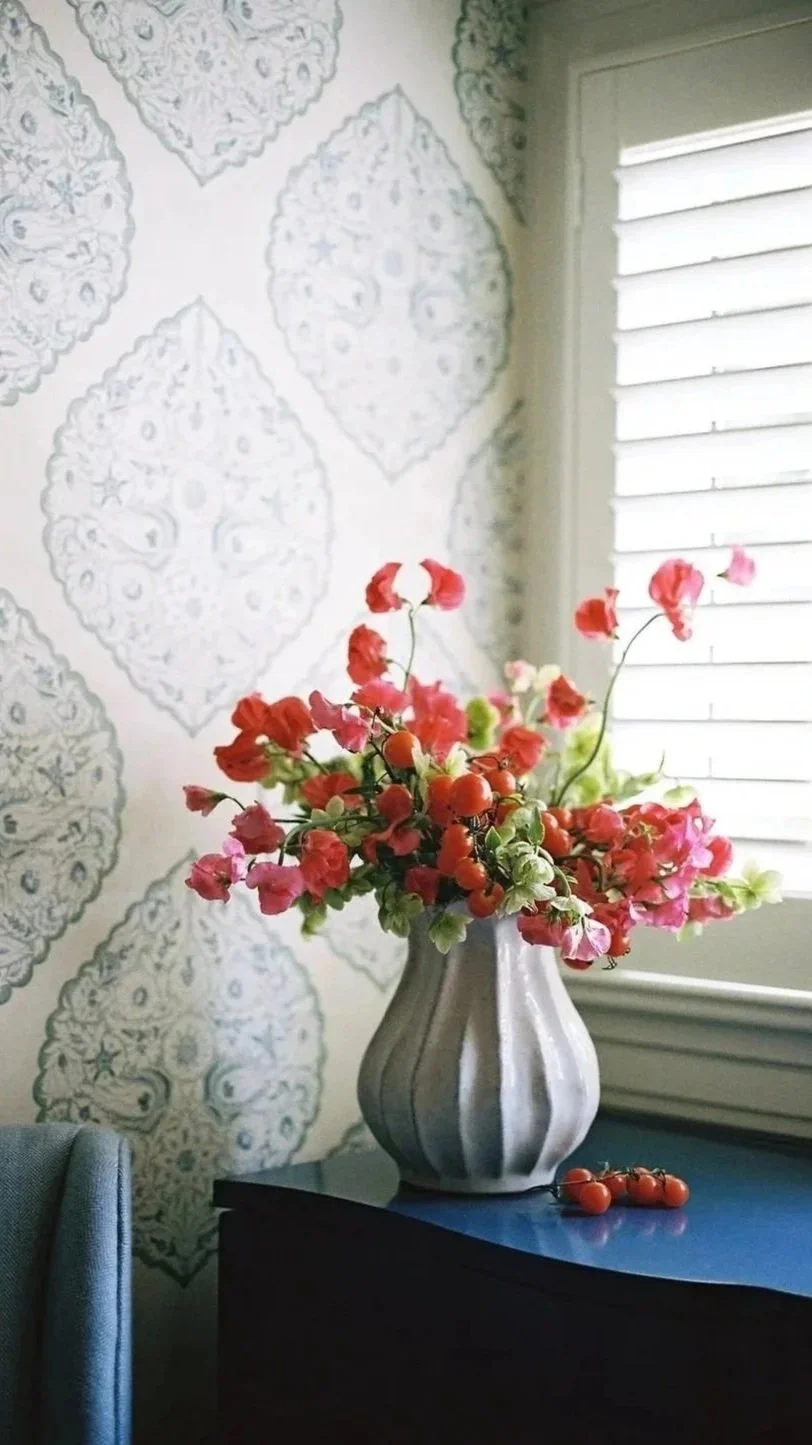 A white ceramic vase with vertical stripes, filled with pink, red, and orange flowers, sits on a dark blue table near a window with white blinds, and a patterned wall background.