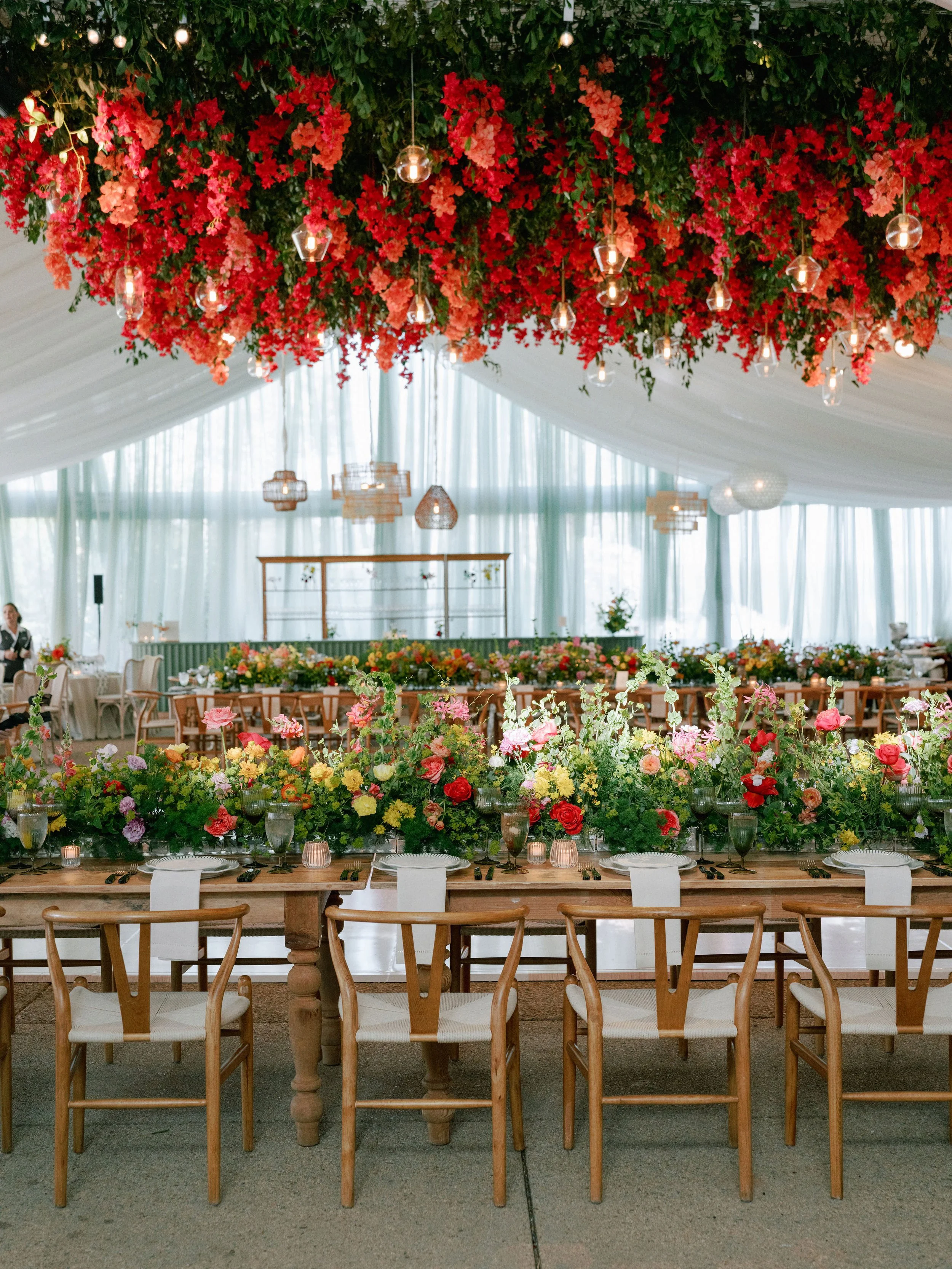A decorated banquet setting with a long wooden table adorned with flowers, candles, and glassware. Overhead, a floral installation with pink and red flowers and hanging lights creates a festive atmosphere. In the background, white drapes and addition