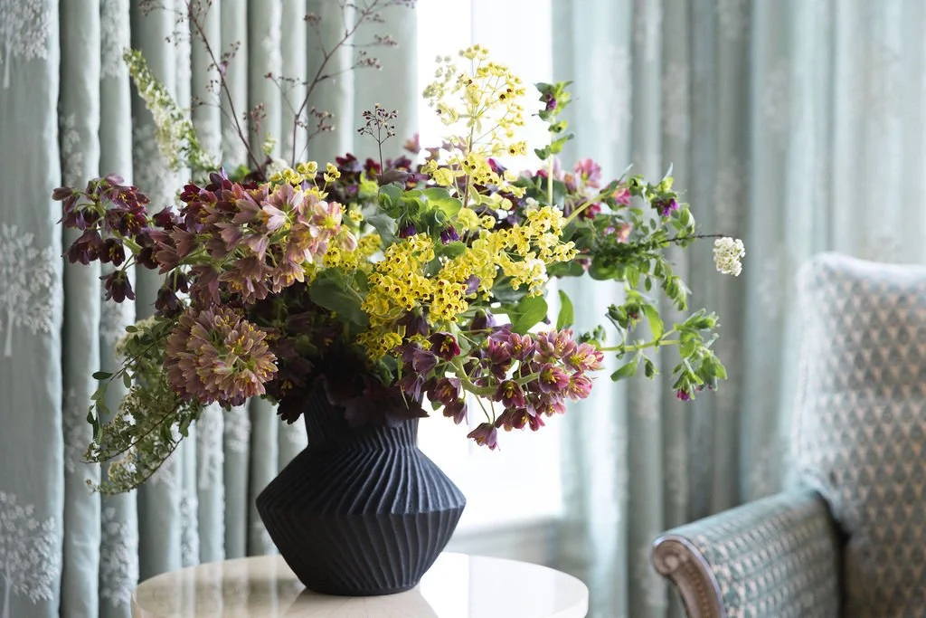 A large bouquet of colorful flowers in a black geometric vase placed on a white table, with curtains and a patterned armchair in the background.