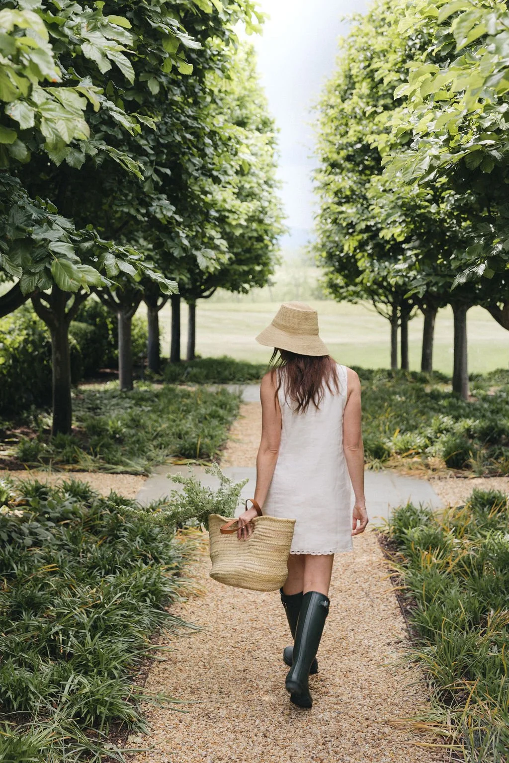 A woman walking down a gravel path in a garden, carrying a straw basket and wearing a wide-brimmed straw hat, a white sleeveless dress, and black rain boots, surrounded by lush green trees and plants.