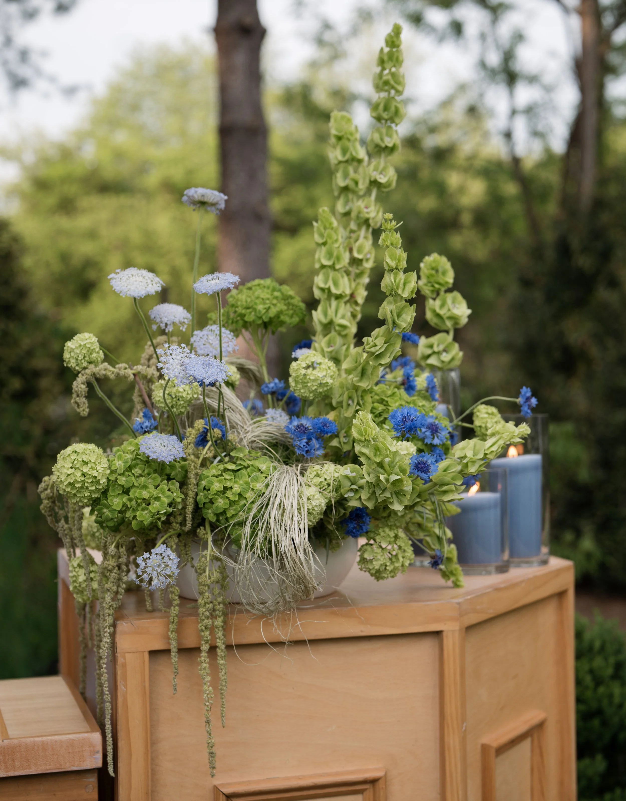 A floral arrangement featuring green, white, and blue flowers placed on a wooden table outdoors with trees in the background.