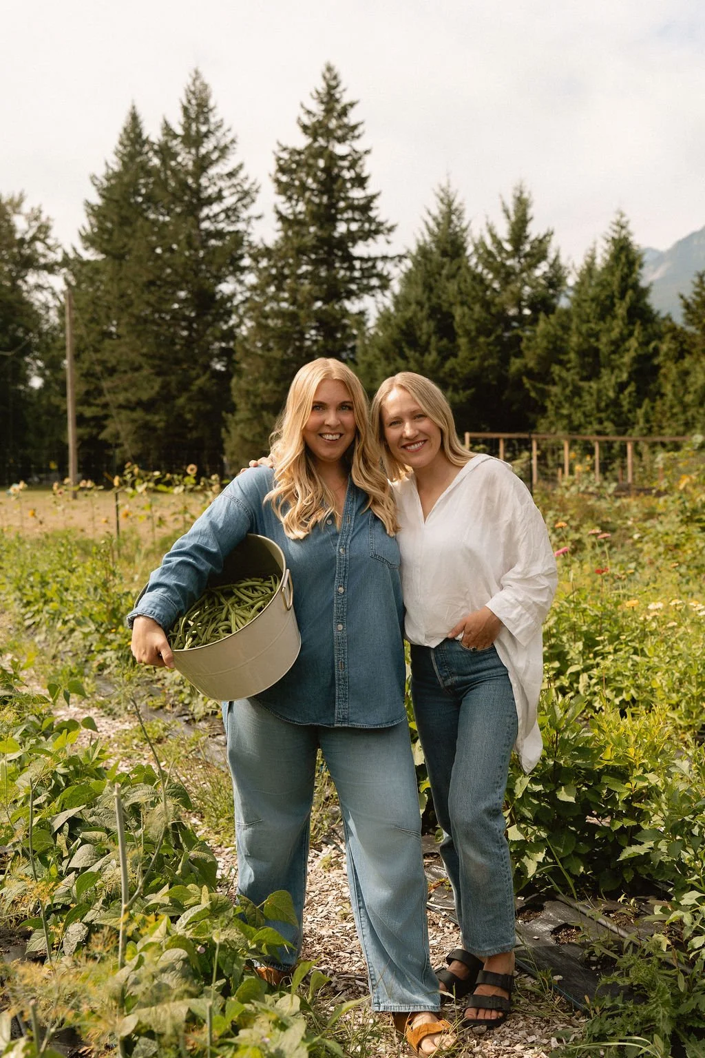 Two women smiling and standing together in a garden or farm, with one holding a bucket of green beans, surrounded by green plants and trees in the background.