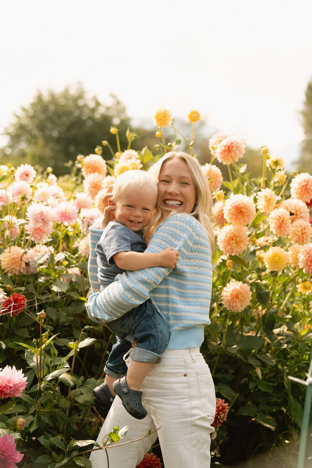 A woman in a blue striped sweater holding a smiling young boy in front of a garden of pink and yellow dahlias.