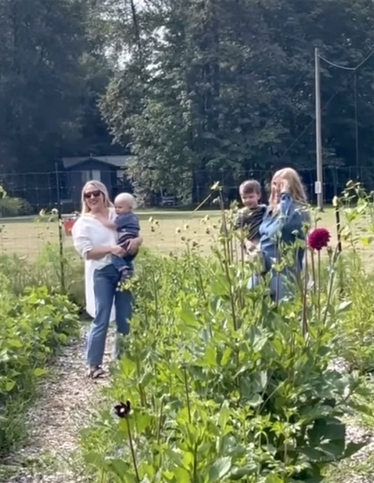 A woman with blonde hair wearing sunglasses and a white shirt is holding a baby. Two children, a boy and a girl, are standing nearby in a garden filled with tall flowers and green plants. The girl is covering her ears, and the boy is smiling. The background includes a grassy field, trees, and a fence.