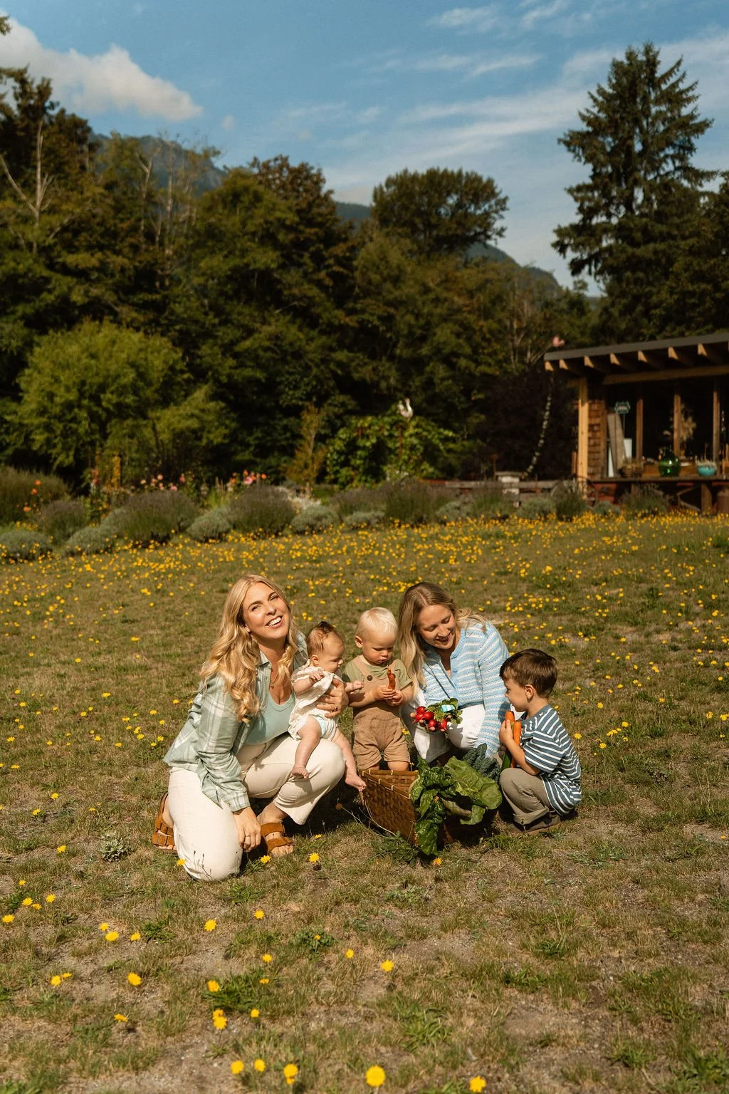 Two women and three children gathering vegetables in a field with yellow flowers, trees, mountains, and a wooden building in the background on a sunny day.