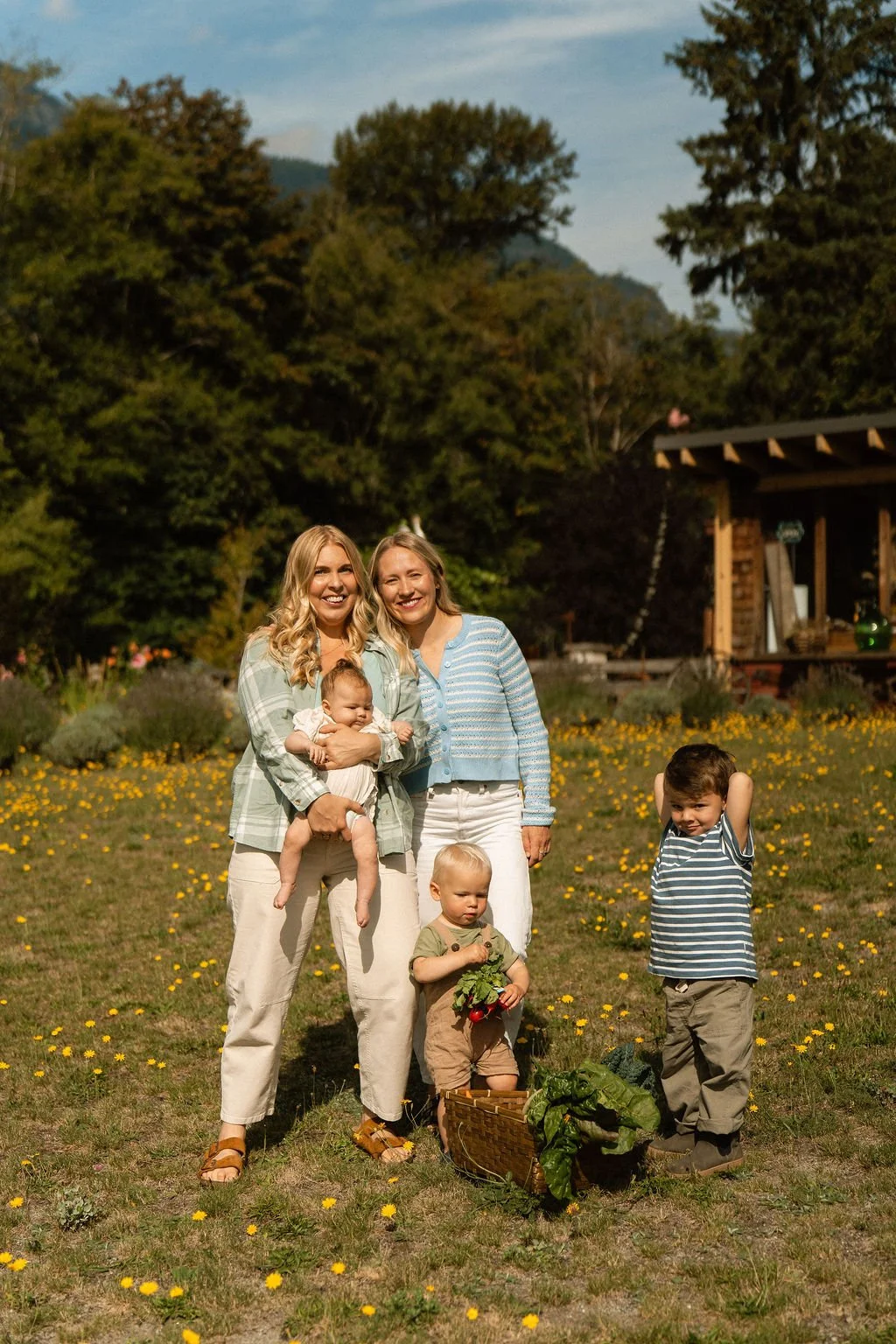 A group of five people, including two adult women and three children, standing outdoors in a field with yellow flowers. The women are smiling, and one is holding a baby. The children are standing nearby, with one holding vegetables and a basket of greens on the ground. There is greenery, trees, and a small building in the background.