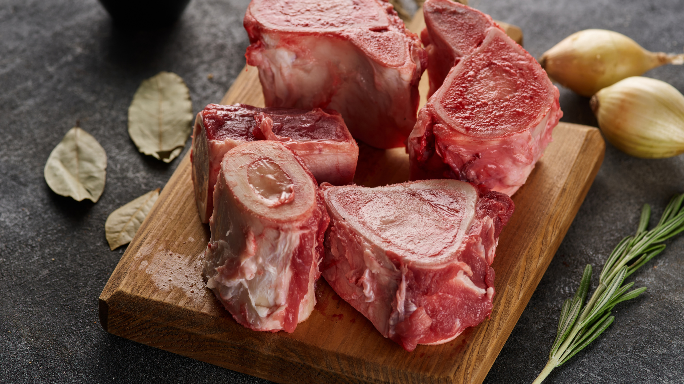 Raw beef bones on a wooden cutting board with bay leaves and rosemary around them.