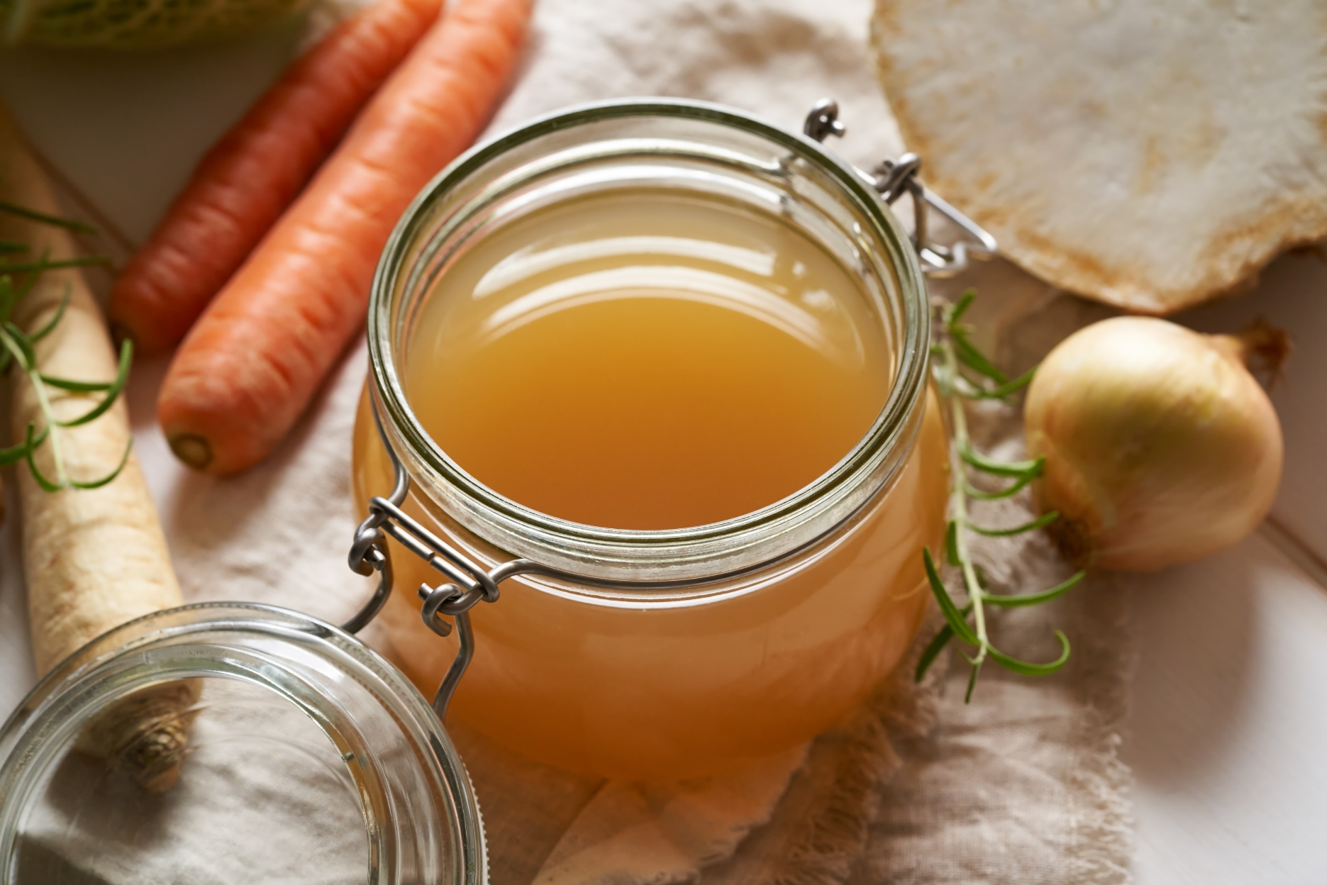 A glass jar filled with honey, surrounded by carrots, garlic, fresh rosemary, and a piece of bread on a white cloth.