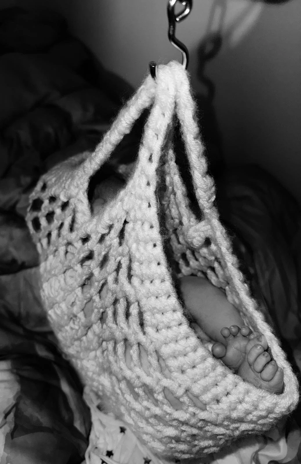 A black-and-white photograph of a small baby's feet in a crocheted blanket, with the blanket hanging from a hook.